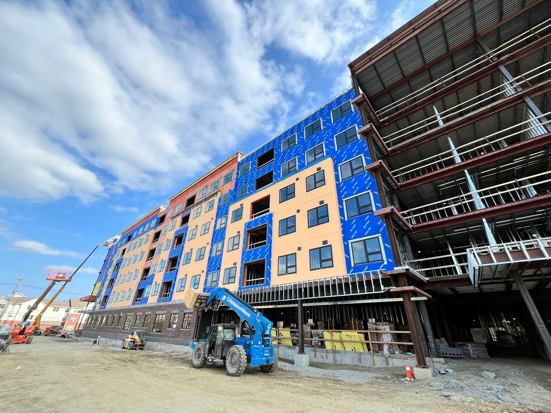 Construction site with a multi-story building, blue tarp scaffolding, and an excavator under a cloudy sky.