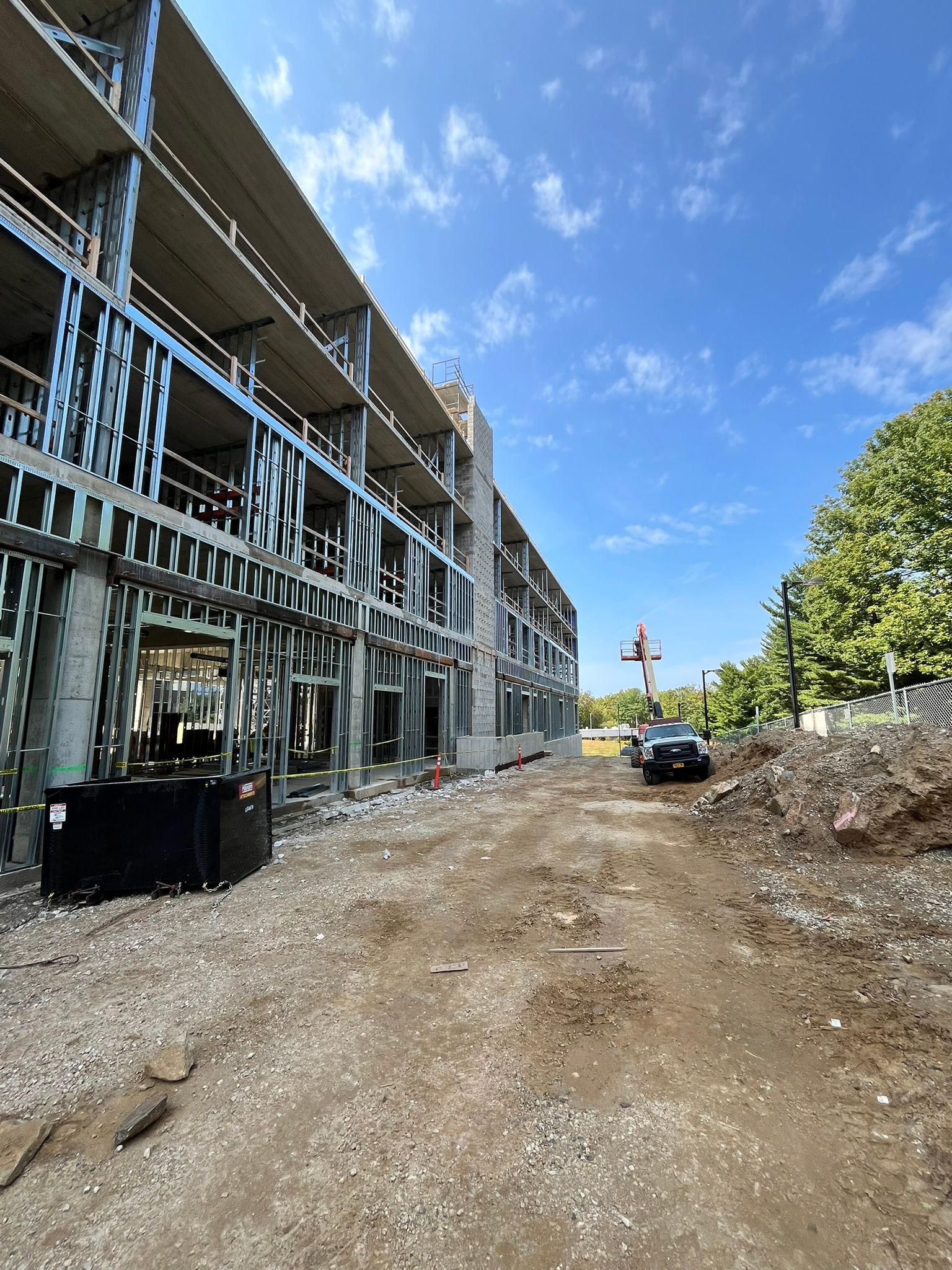 Construction site beside a partially built multi-story building, with a dirt road, parked car, and blue sky.