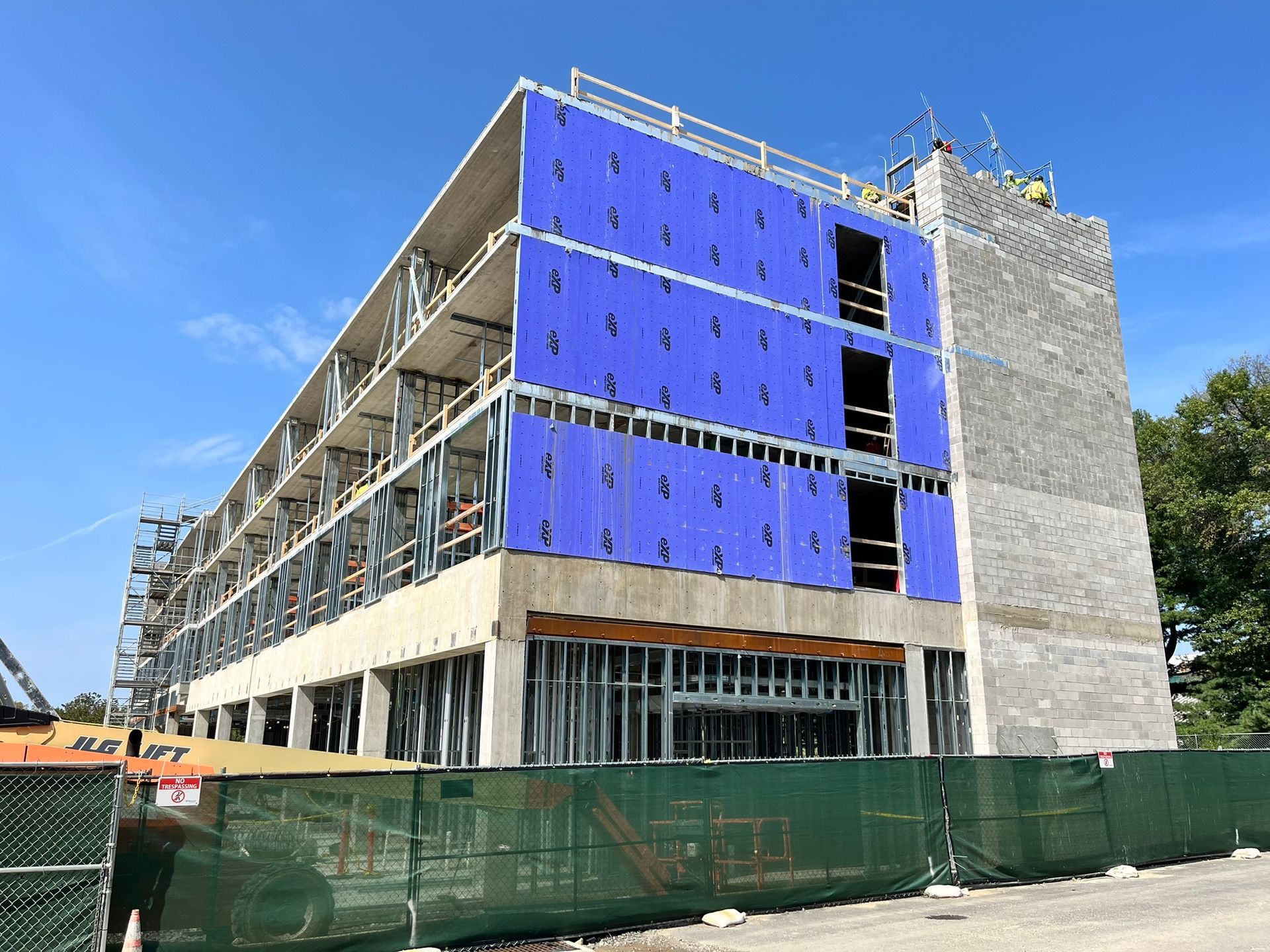 Partially built multi-story building with blue sheathing, scaffolding, and green fencing under a clear sky.