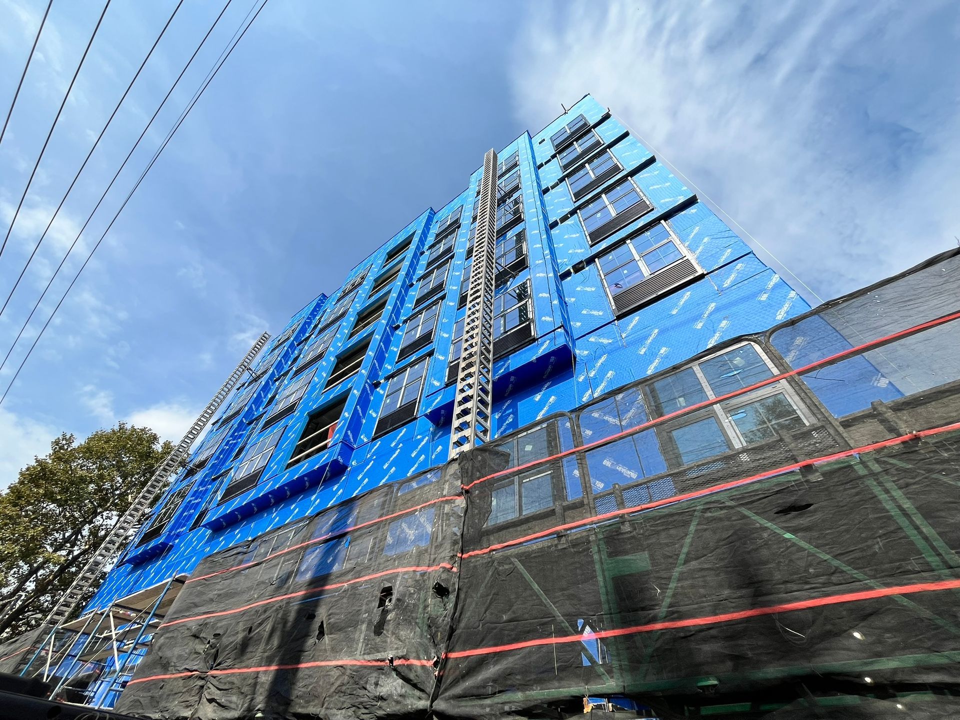 Low-angle view of a tall blue high-rise building against a cloudy sky, with power lines and scaffolding below.