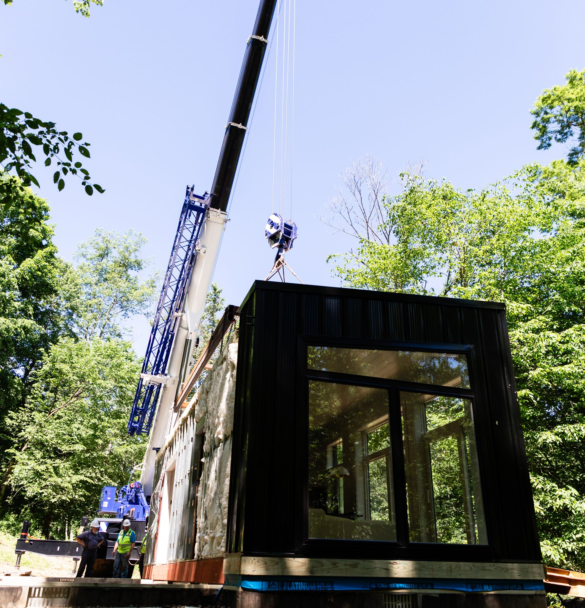 Crane lifting a black modular cabin among trees on a sunny day.