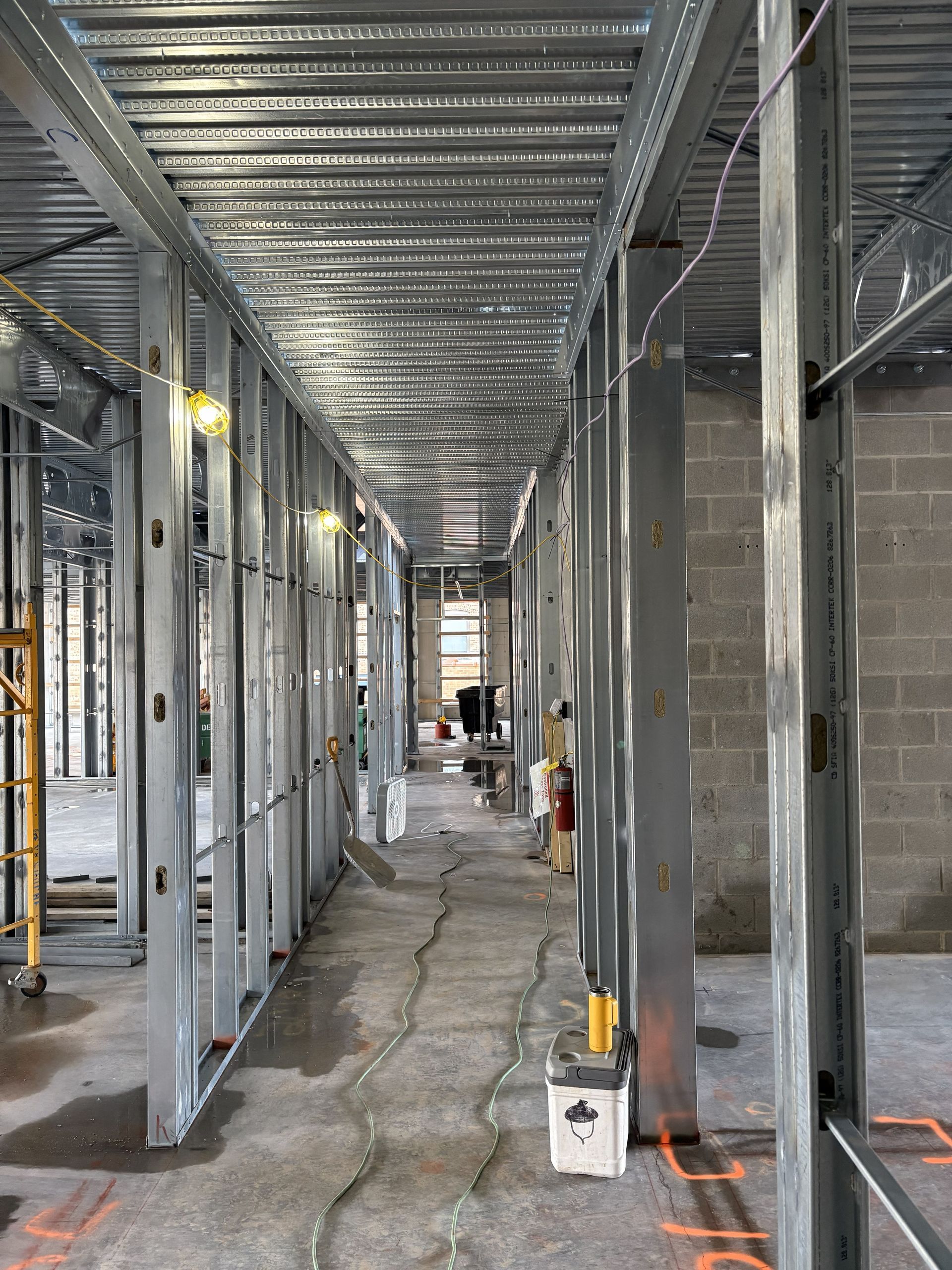 Interior construction site with steel framing, exposed studs, and unfinished floor in a hallway-like space.