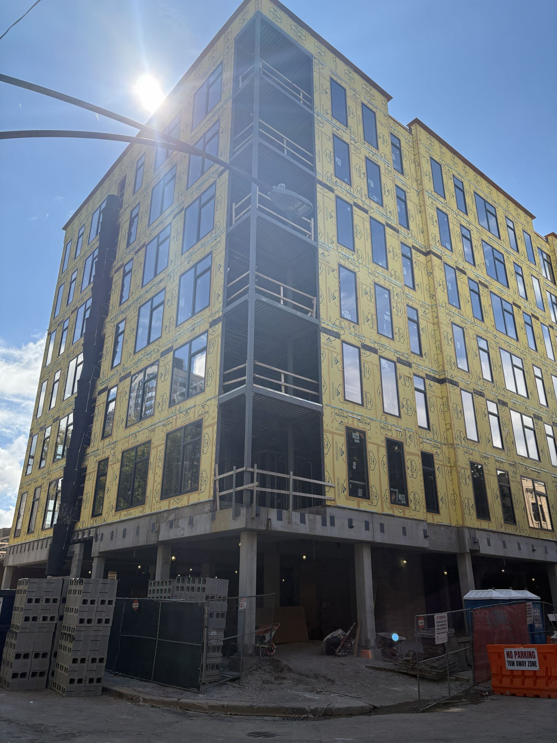 Sunlit yellow-and-black multi-story building under construction on a city street, with scaffolding and debris below.