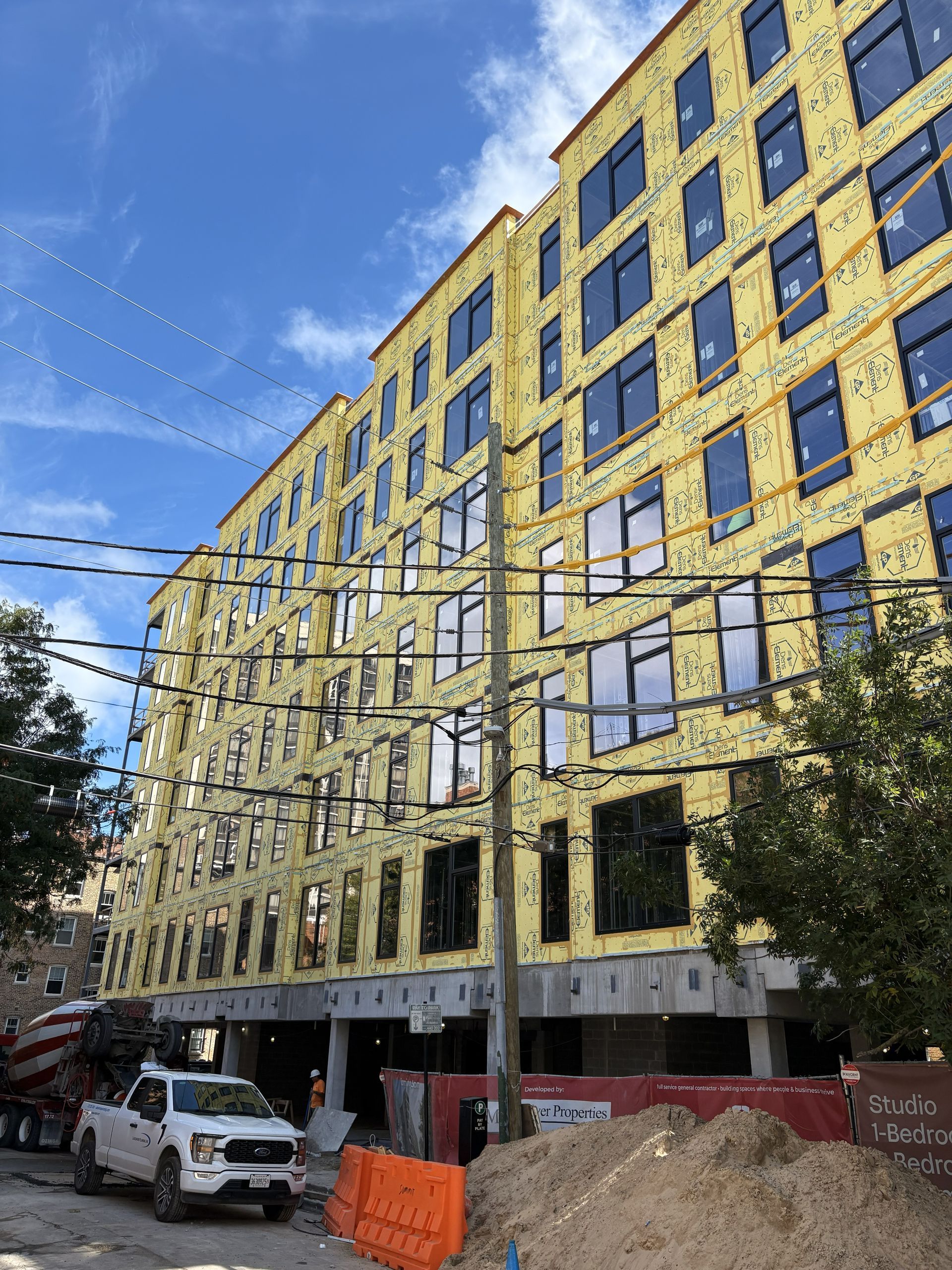 Tall yellow multi-story building under construction with workers’ vehicles and a blue sky.