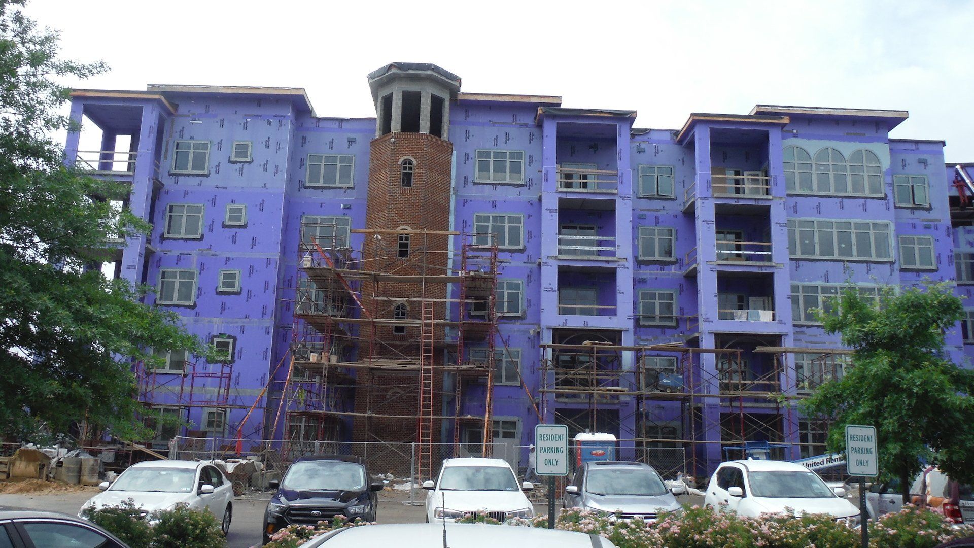 A large building with purple insulation panels under construction with cars parked in front of it.