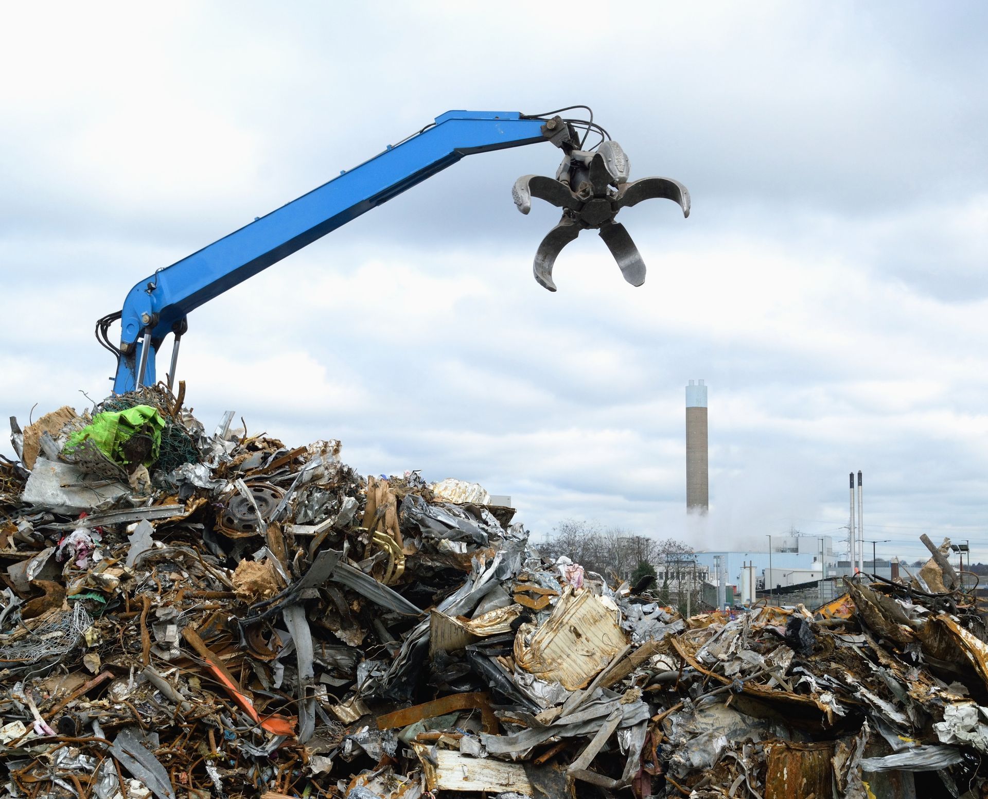 Blue crane grapple lifting scrap metal above a large pile at a recycling yard.