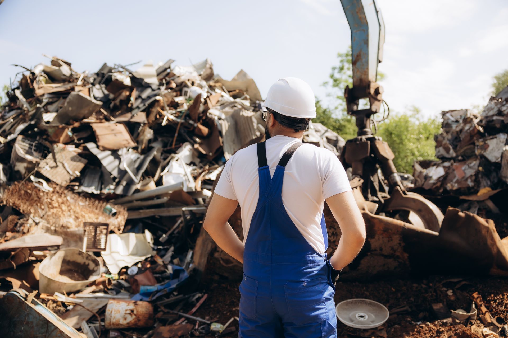 Scrap metal yard worker in hard hat and overalls inspecting recycled metal pile at salvage facility. Scrap metal yard worker in hard hat and overalls inspecting recycled metal pile at salvage facility.