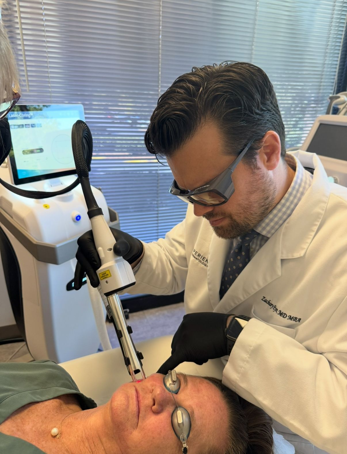A provider in a white coat and protective eyewear uses a laser device on a patient’s face in a medical office setting.