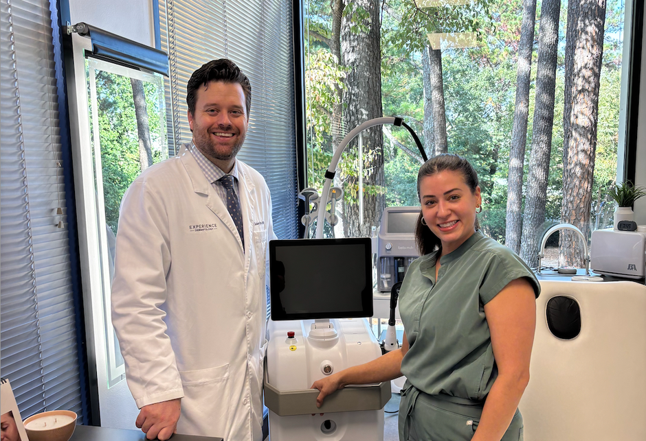 Doctor and clinician smiling beside a medical scanner in a bright office with trees outside the window