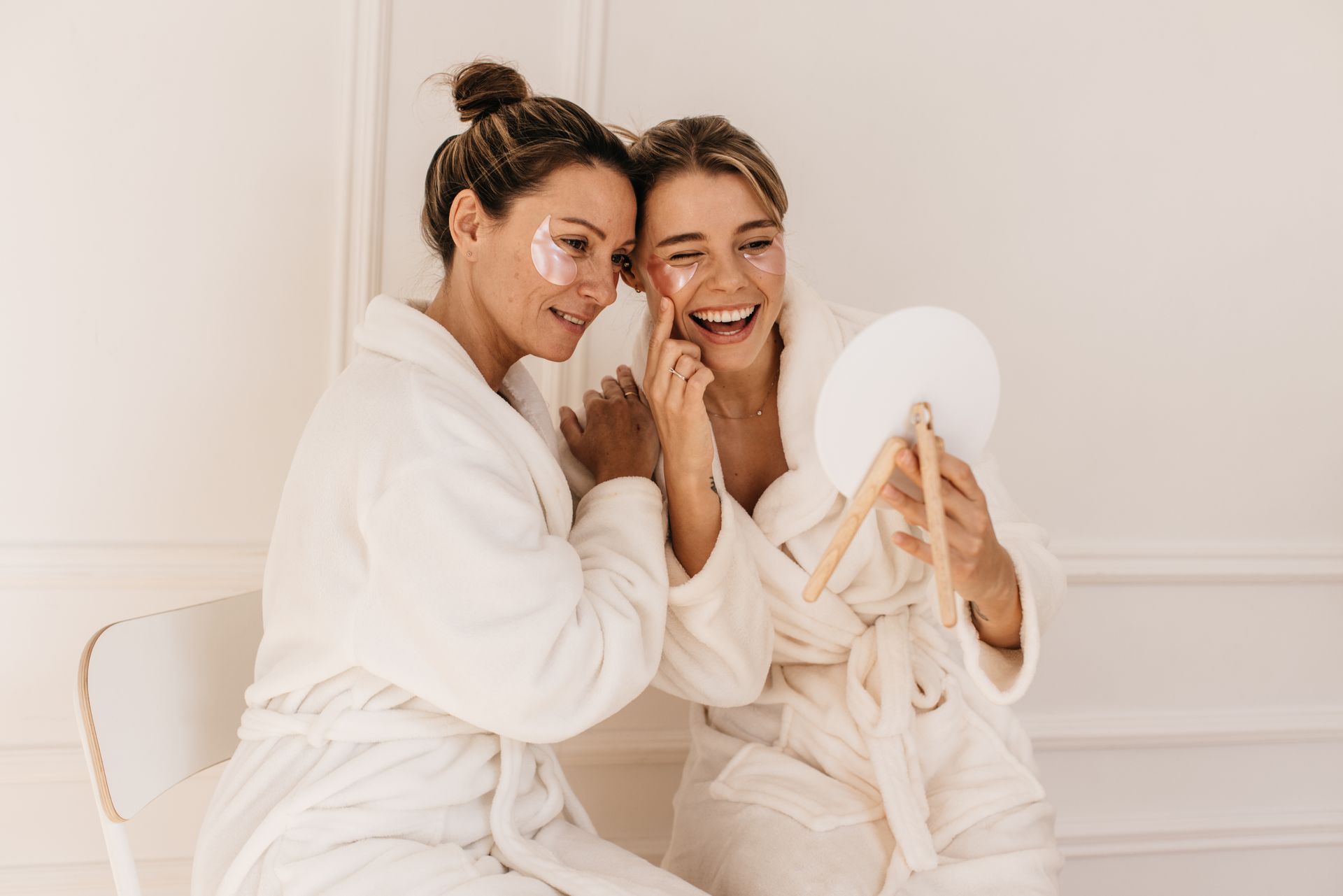 Two people in white robes and under-eye patches laugh together while looking into a small handheld mirror.