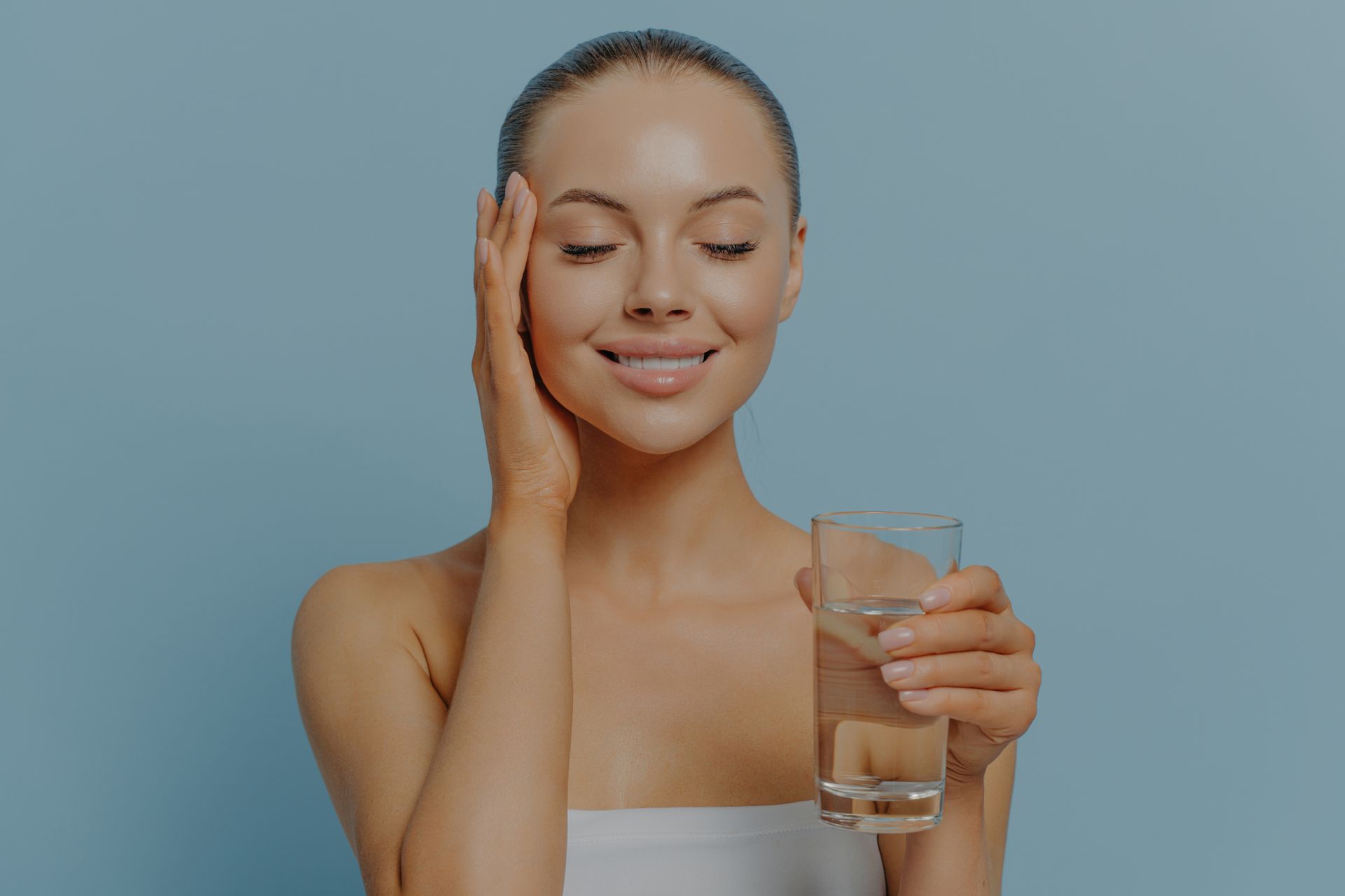 Smiling woman in a white top holding a glass of water against a blue background