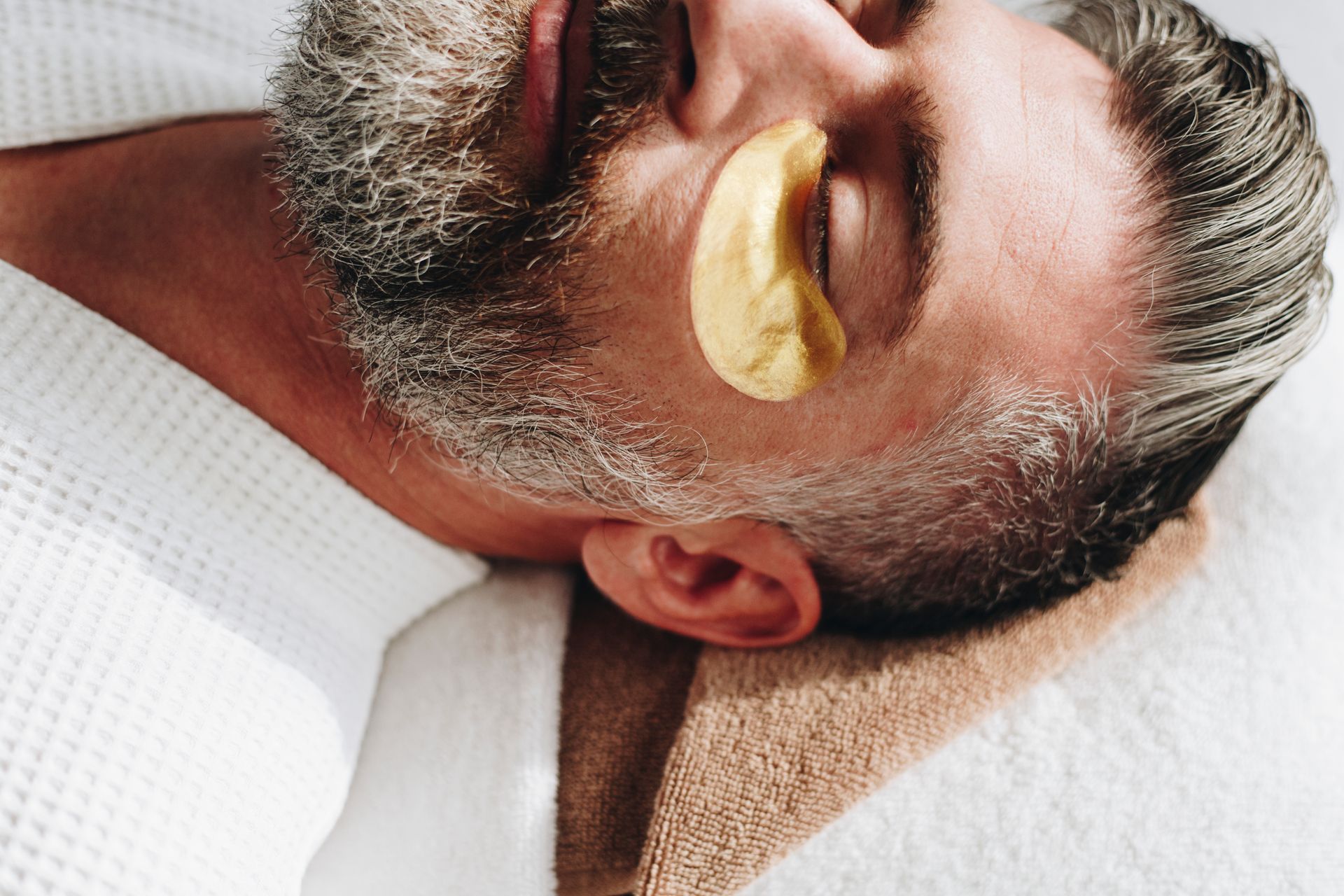 A person with a beard lies on a towel wearing a gold under-eye patch, eyes closed in a relaxing spa setting.