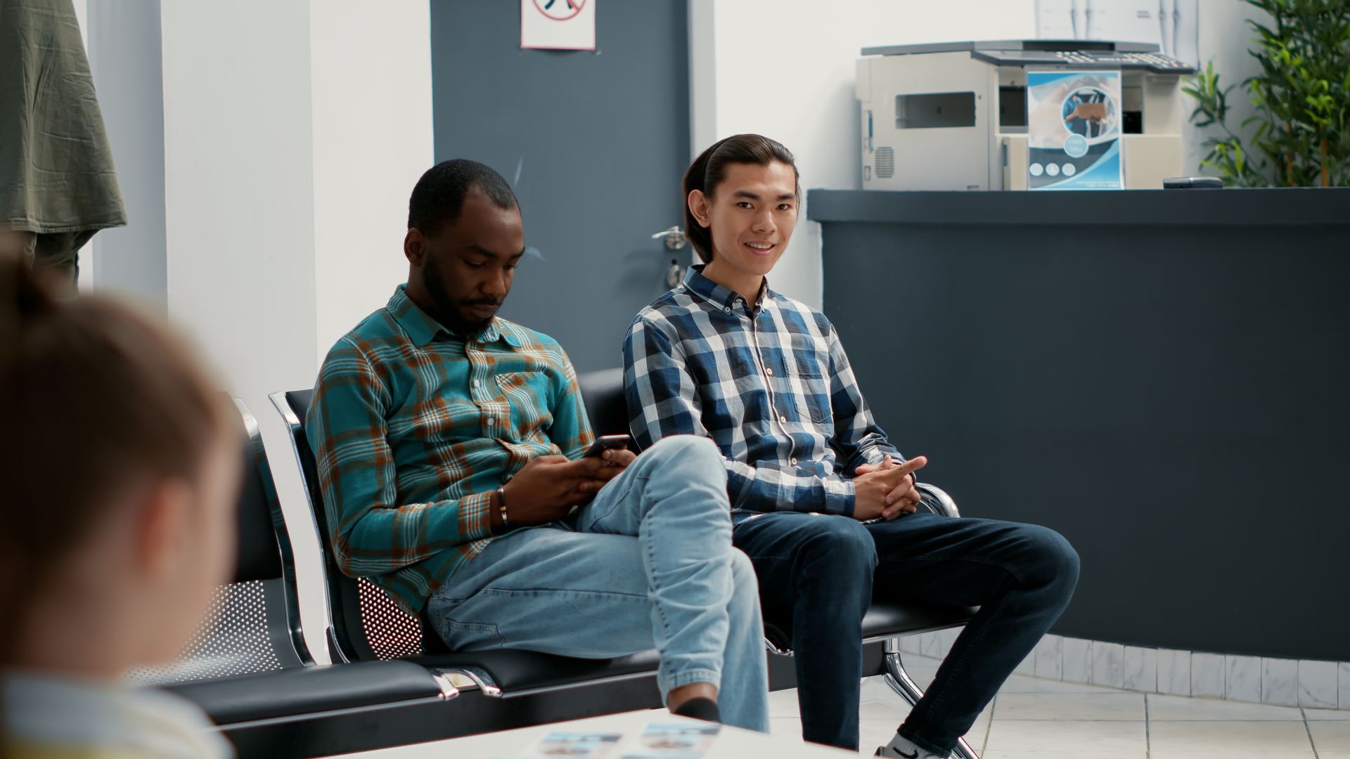 Two men seated in a waiting room, one looking at a phone and the other smiling on a chair.