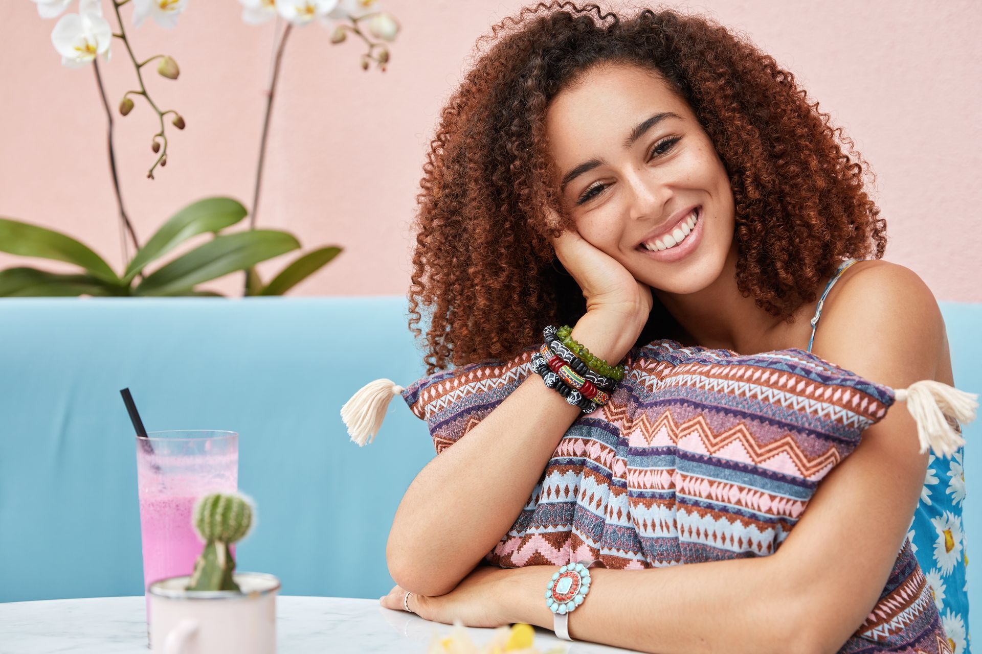 A person with curly hair smiling and resting their chin on a patterned pillow at a table with a pink drink and a cactus.