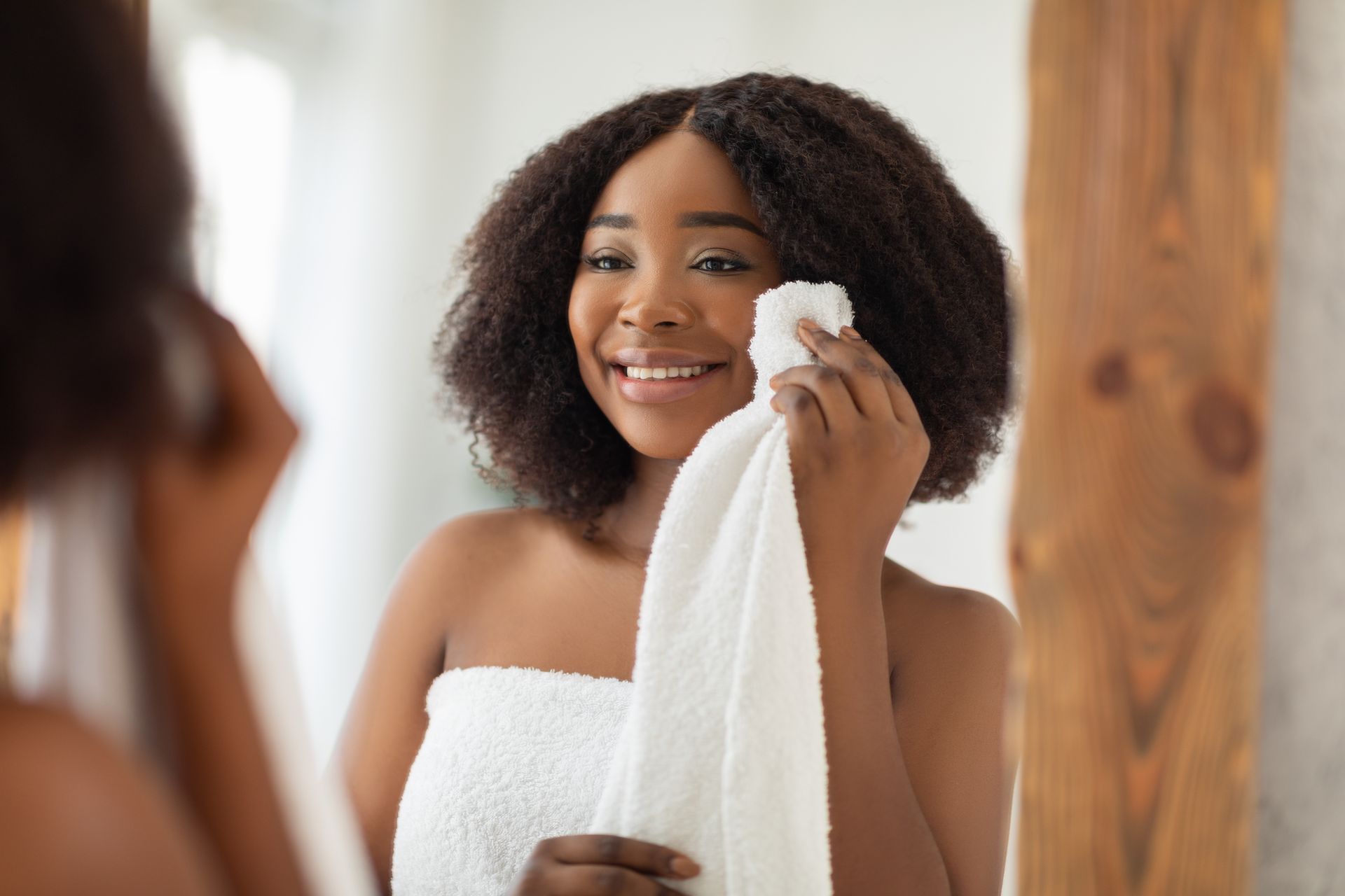 Woman smiling in a towel while drying her face in a bathroom mirror