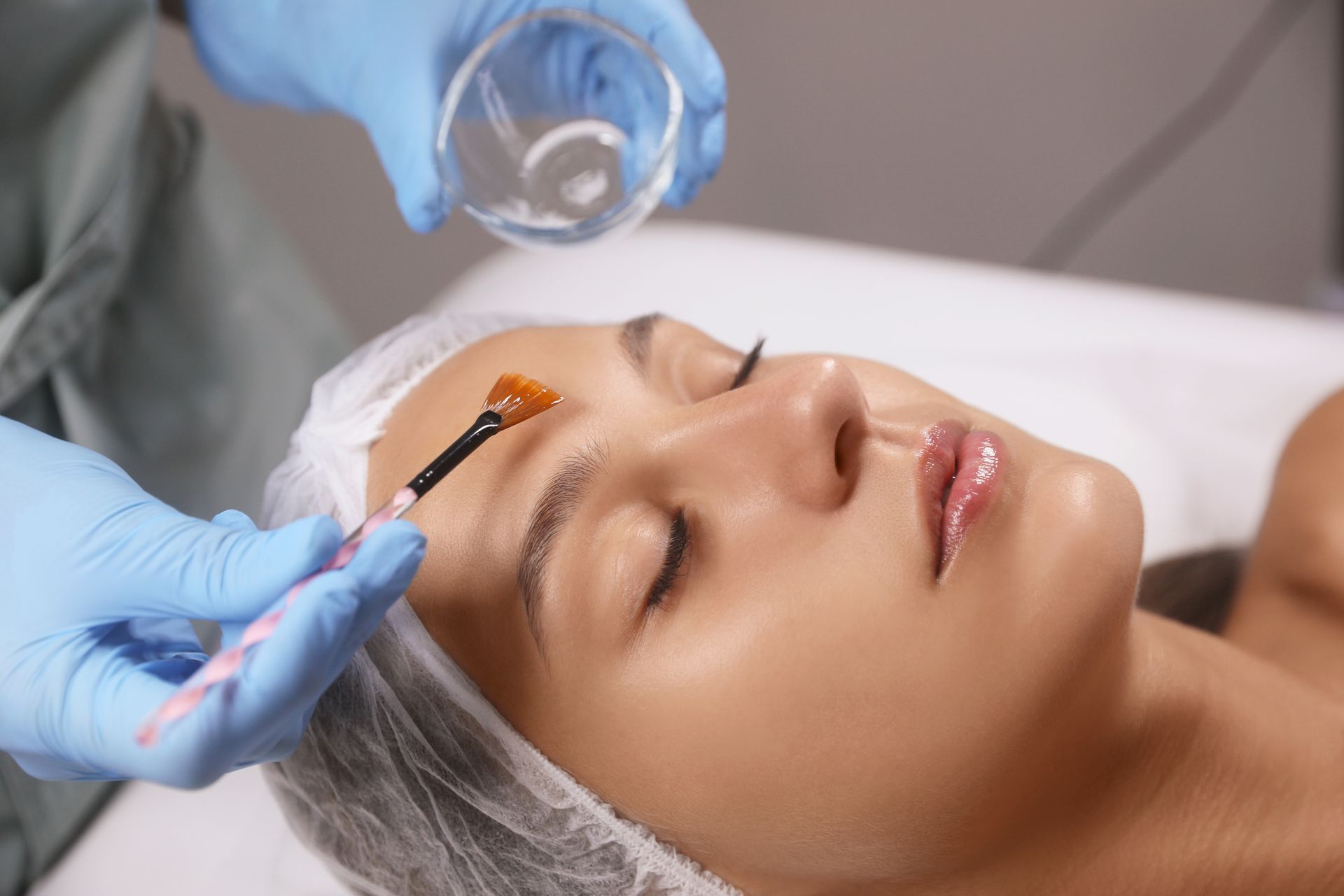 A gloved professional uses a brush to apply a treatment to a person's forehead in a spa setting.
