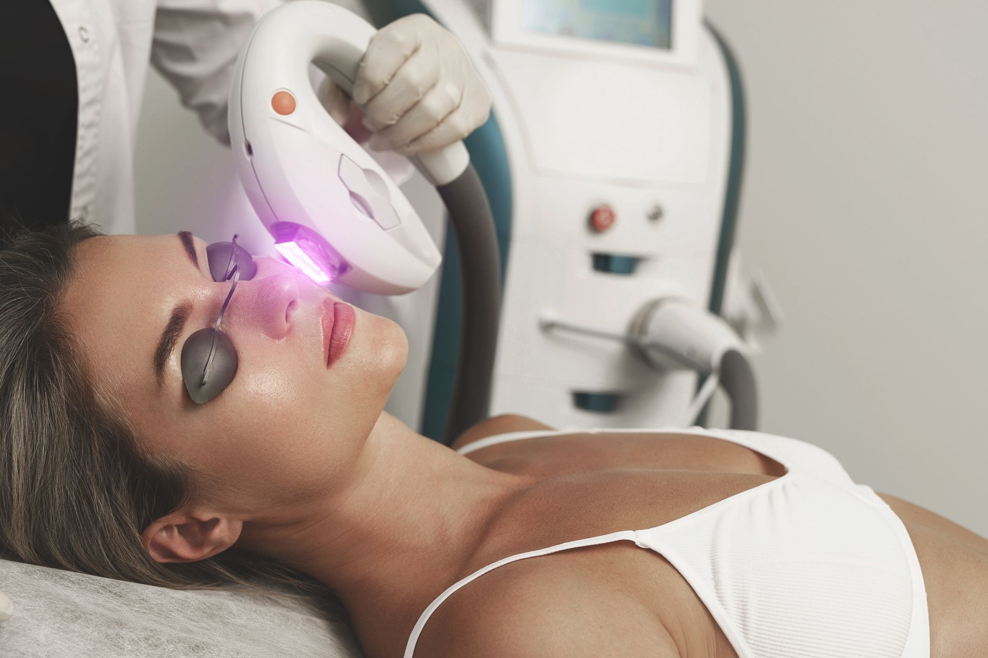 Woman receiving facial laser treatment in a clinic, with a white device emitting pink light on her face
