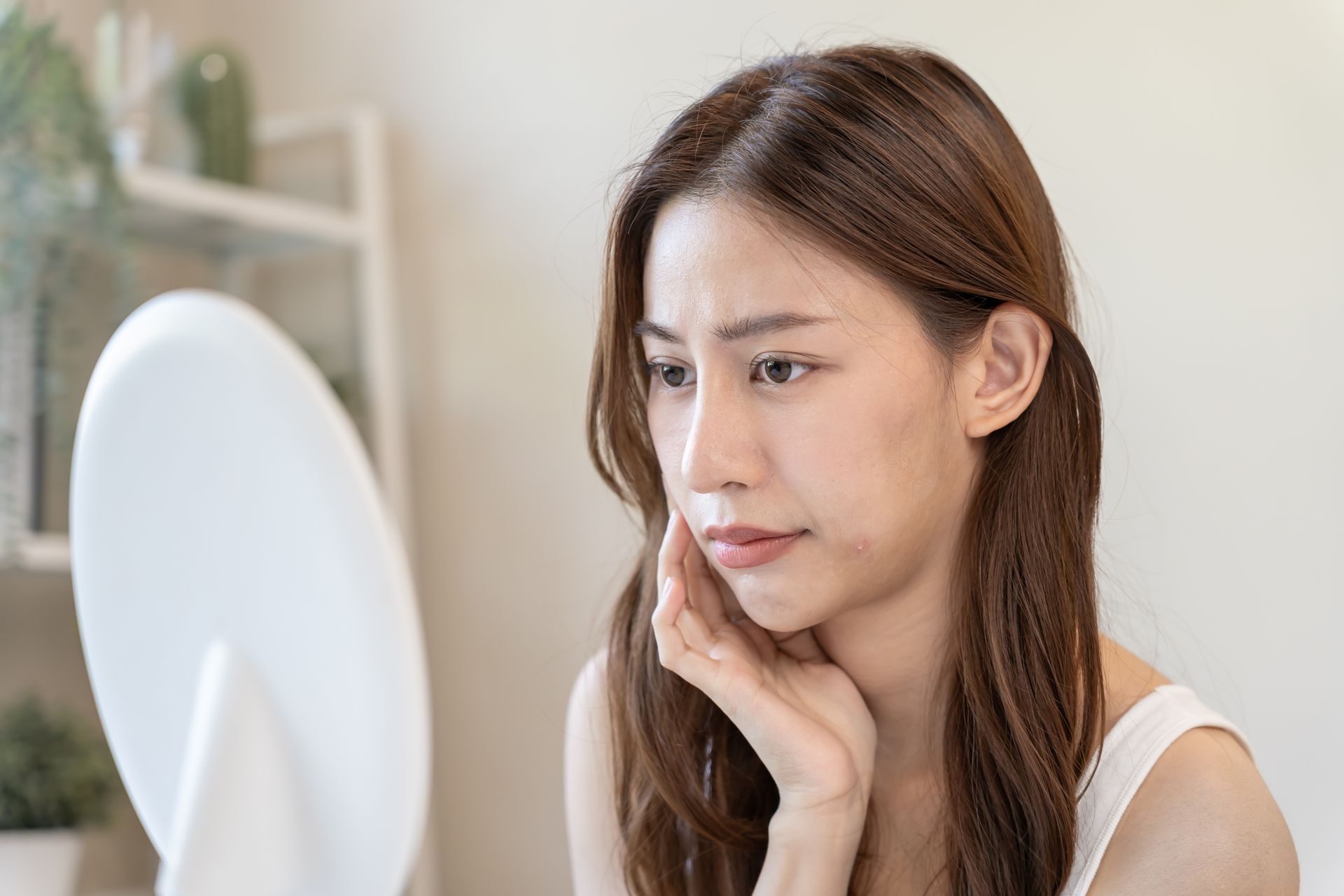 Woman examining her face in a mirror, touching her cheek with a concerned expression.