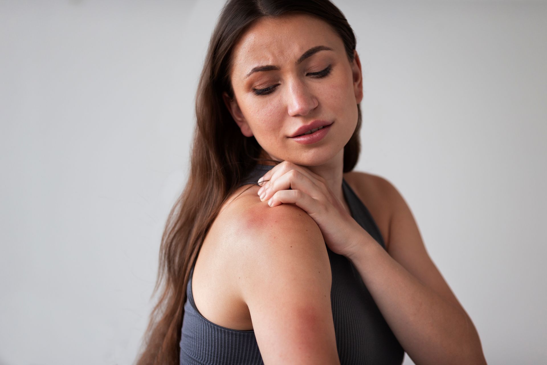 Woman in a gray tank top touching her shoulder with a pained expression against a plain background