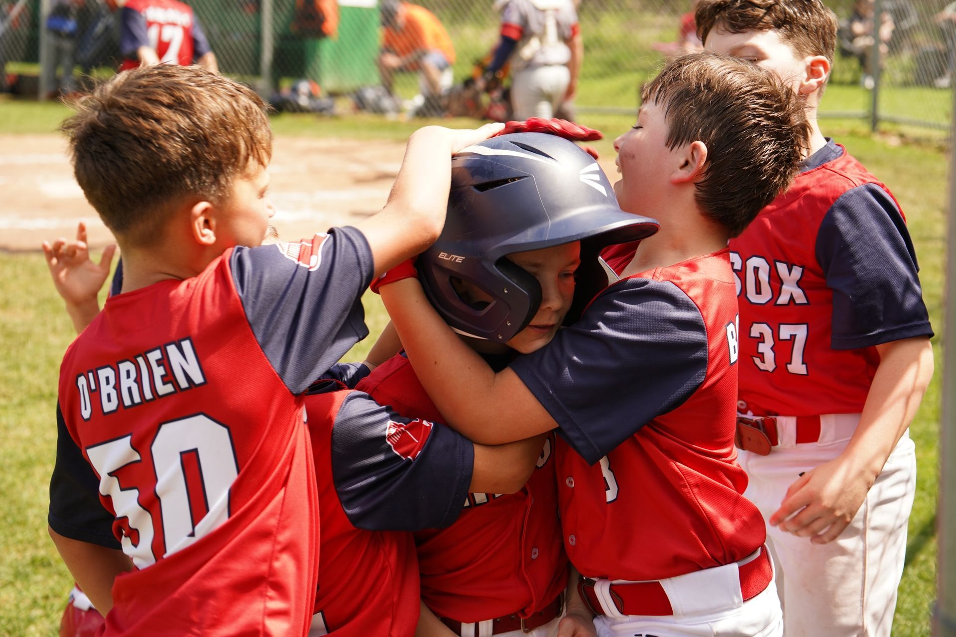 Youth baseball players in red uniforms huddle around a teammate wearing a batting helmet.