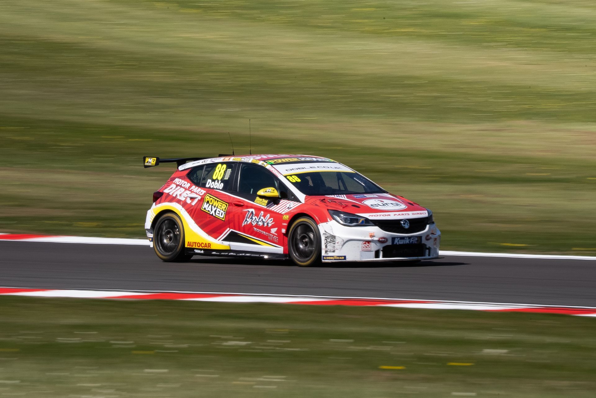 Race car, red and yellow, on track, blurred background of green hills.