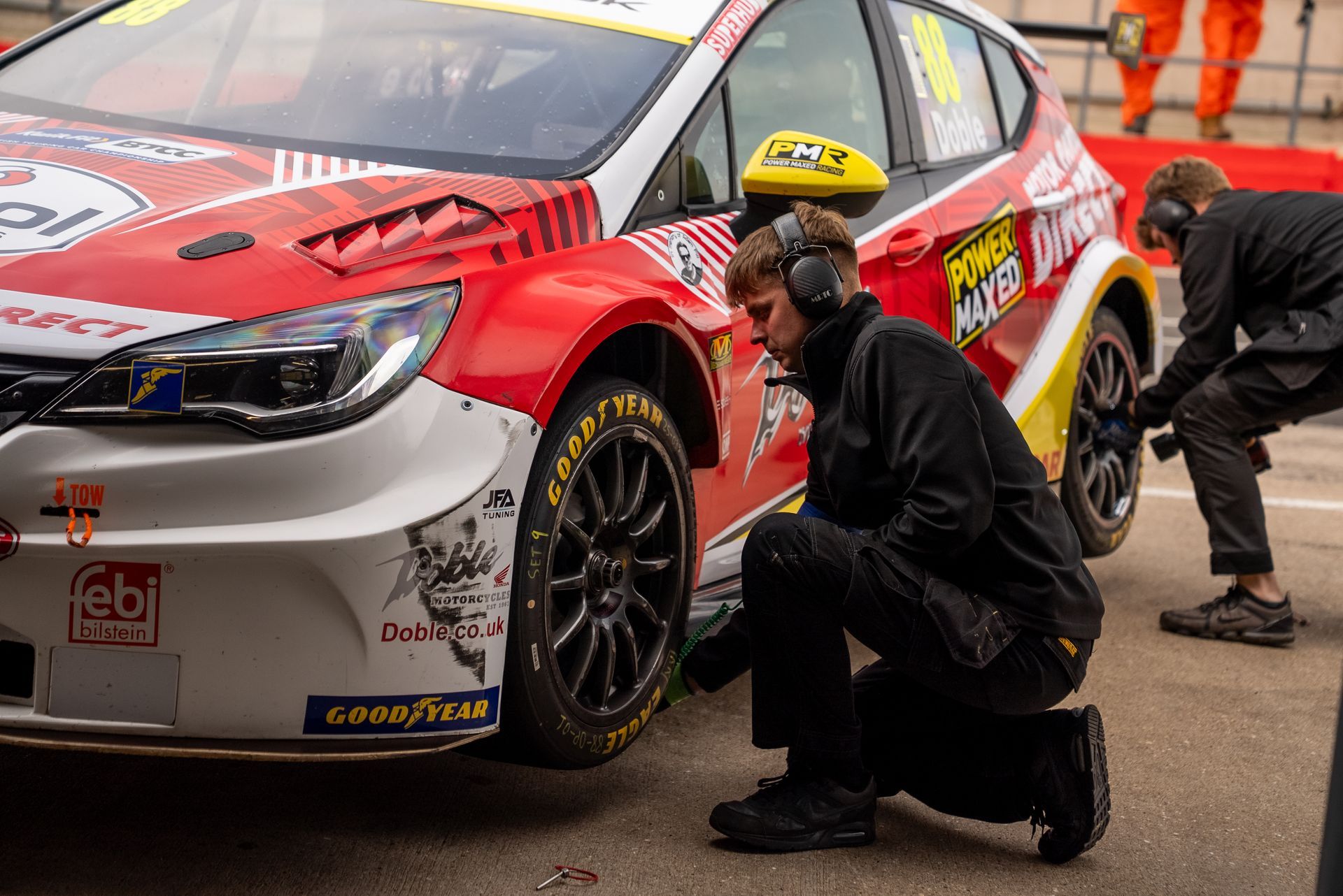 Crew member servicing a red and white race car's tire in a pit lane; another person in the background.