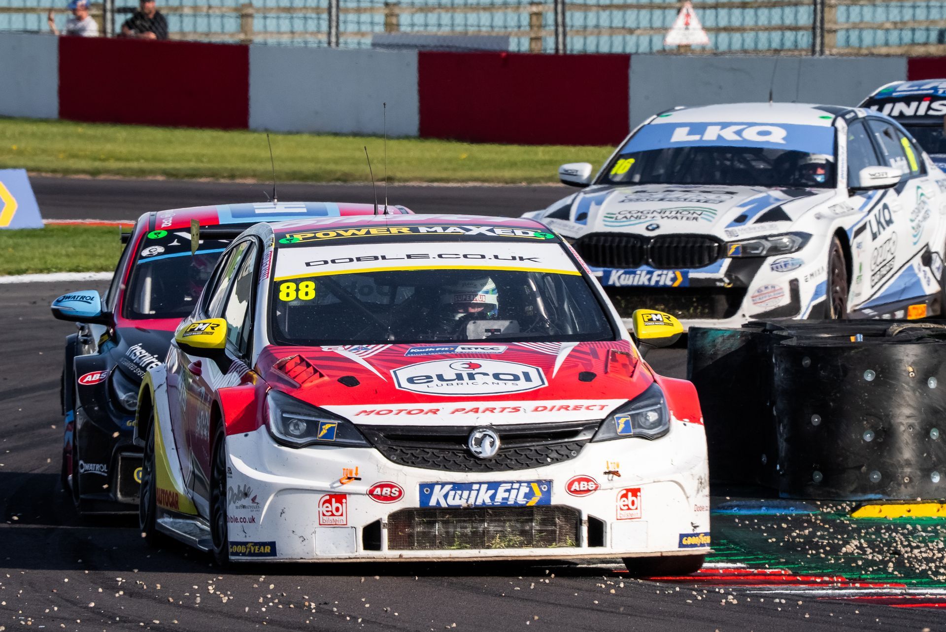 Race cars on a track; the lead car is red and white with sponsor logos, another car in the background is black and red.