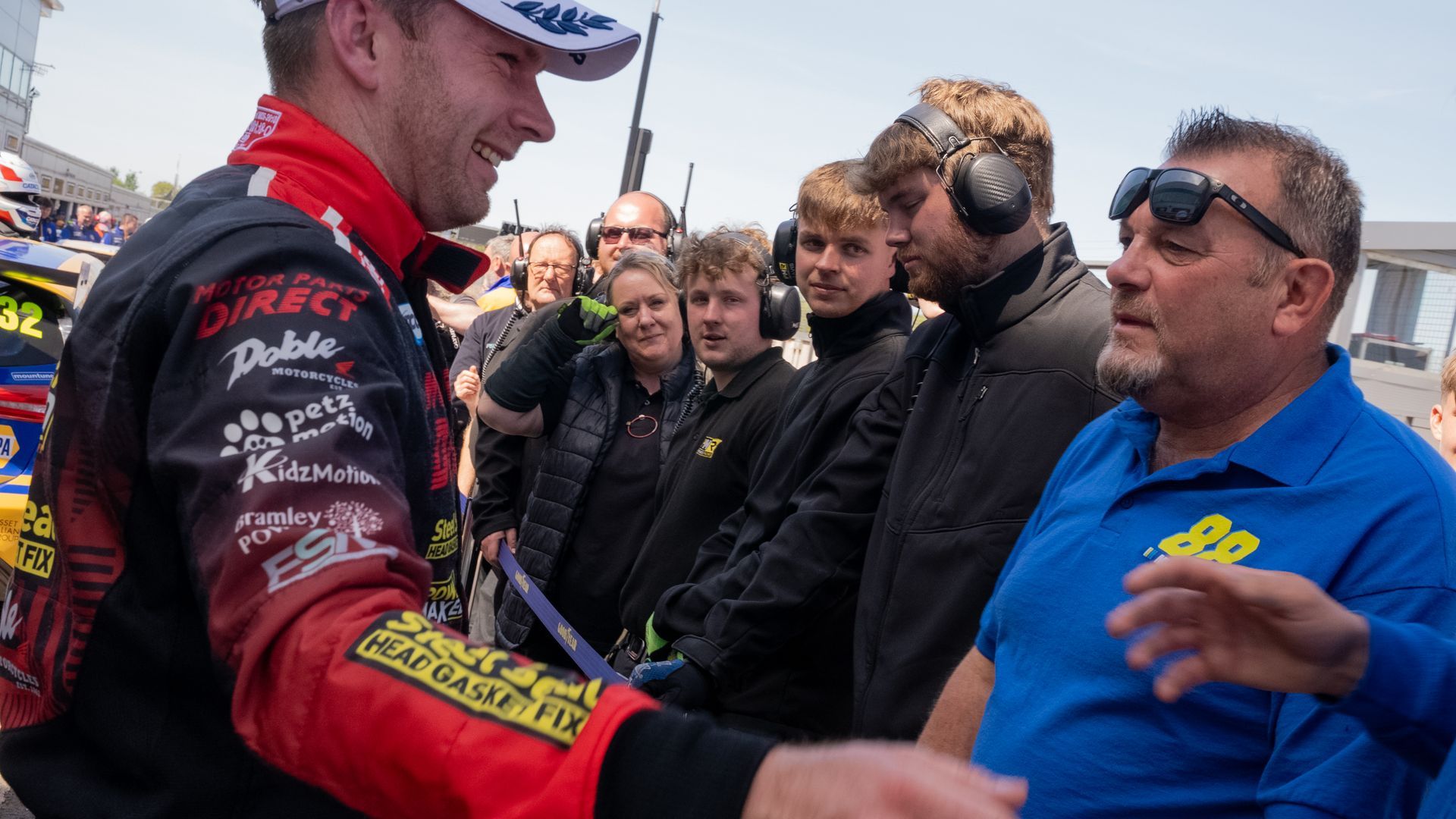 Race car driver smiling, shaking hands with a man in blue, surrounded by crew and onlookers at a racetrack.