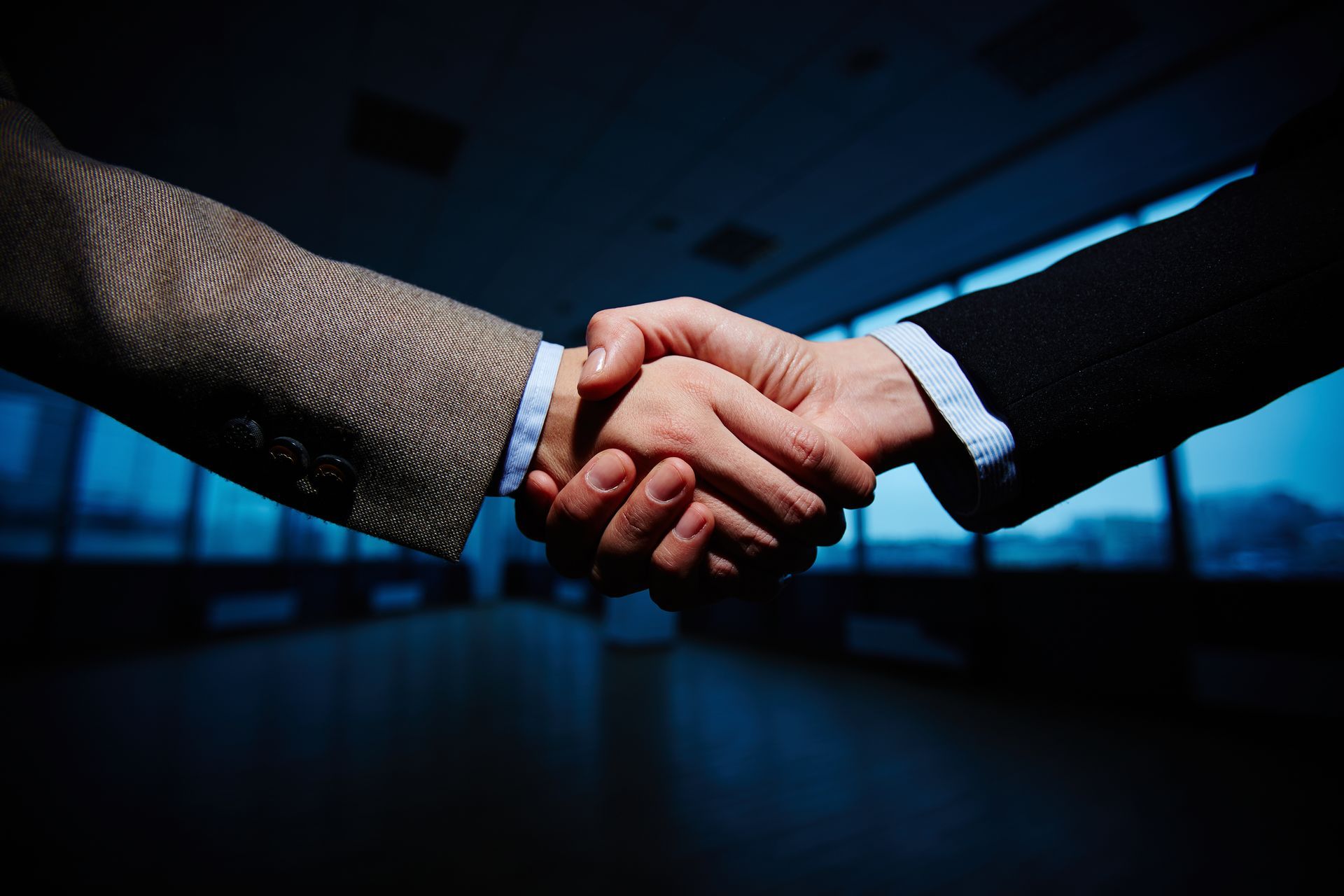 Two people shaking hands in a business setting, dark interior with blue toned windows.