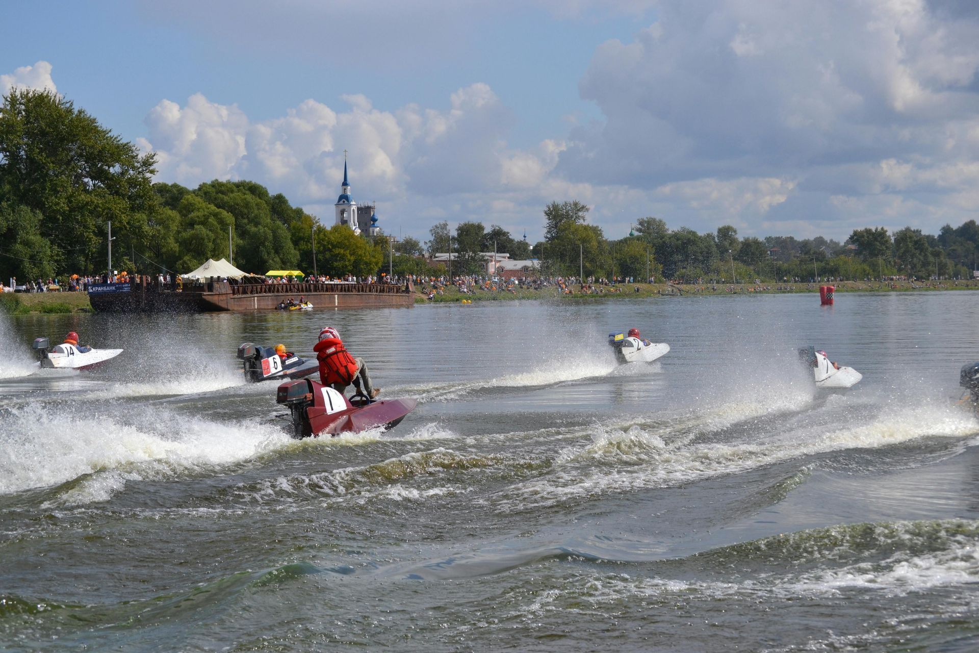 Jet skis racing across a lake, spraying water near a tree-lined shore under a partly cloudy sky