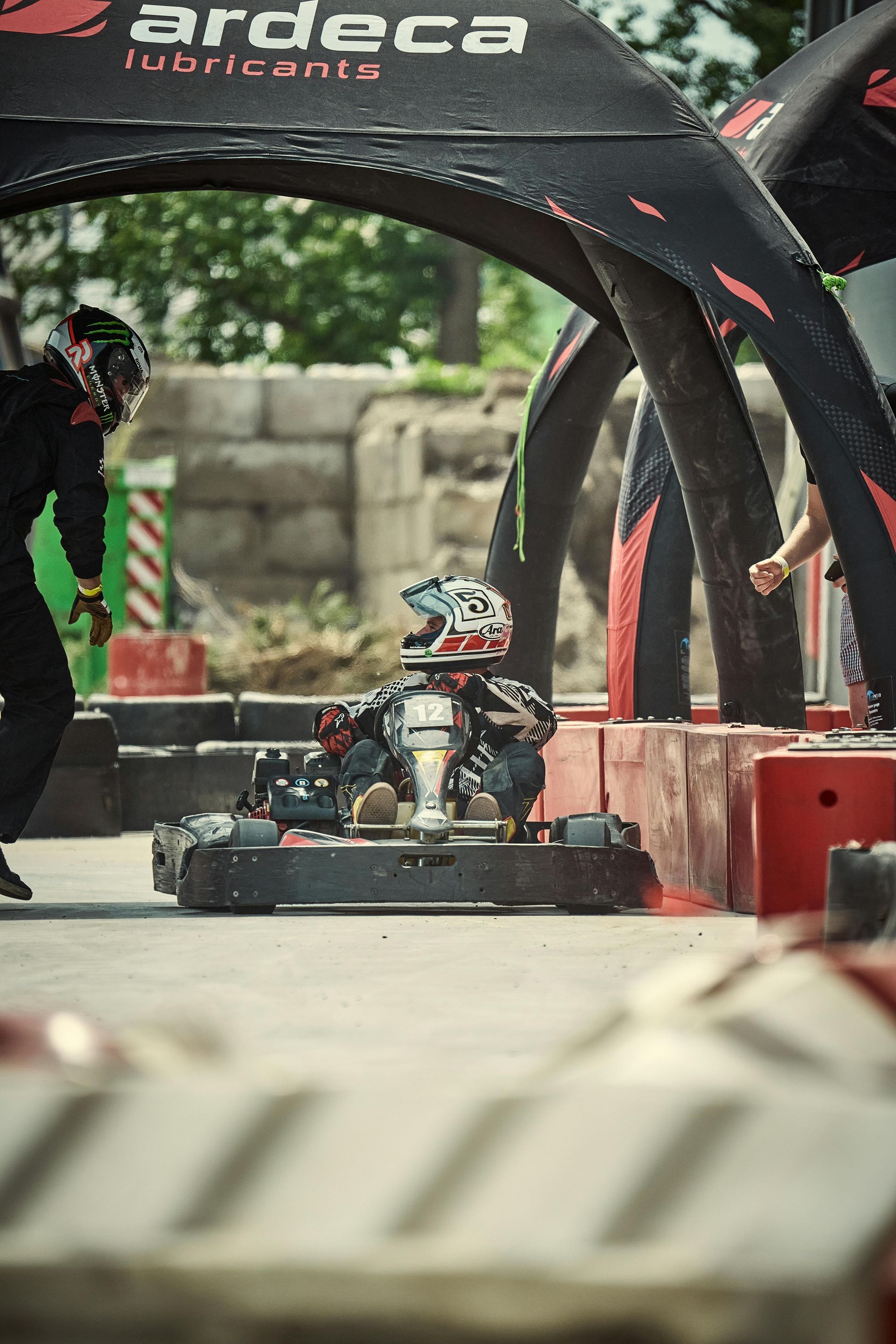 Go-kart racer in helmet and suit at starting line, under archway. Black and red barriers in a paved outdoor setting.