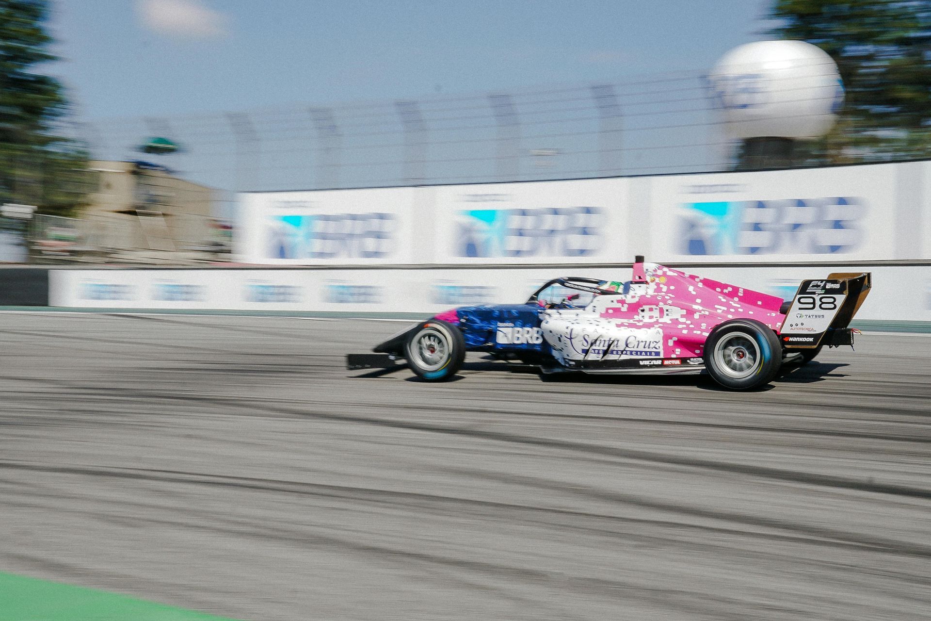 Race car, blue and pink, on a racetrack with sponsor signs in the background.