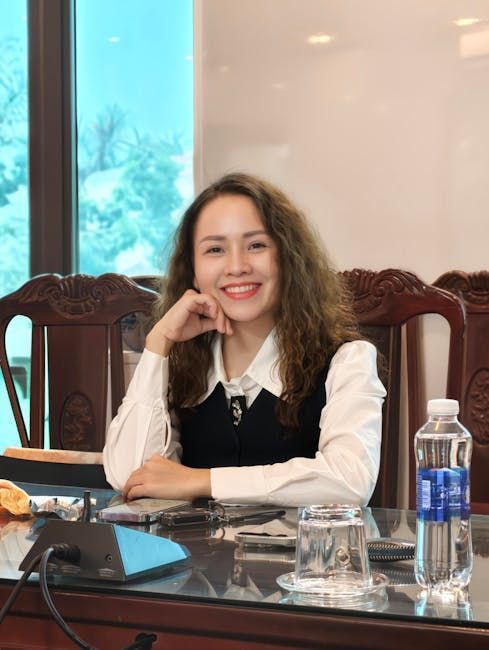 Woman smiling, seated at a desk, wearing a white shirt and black vest, with water bottle and glass.