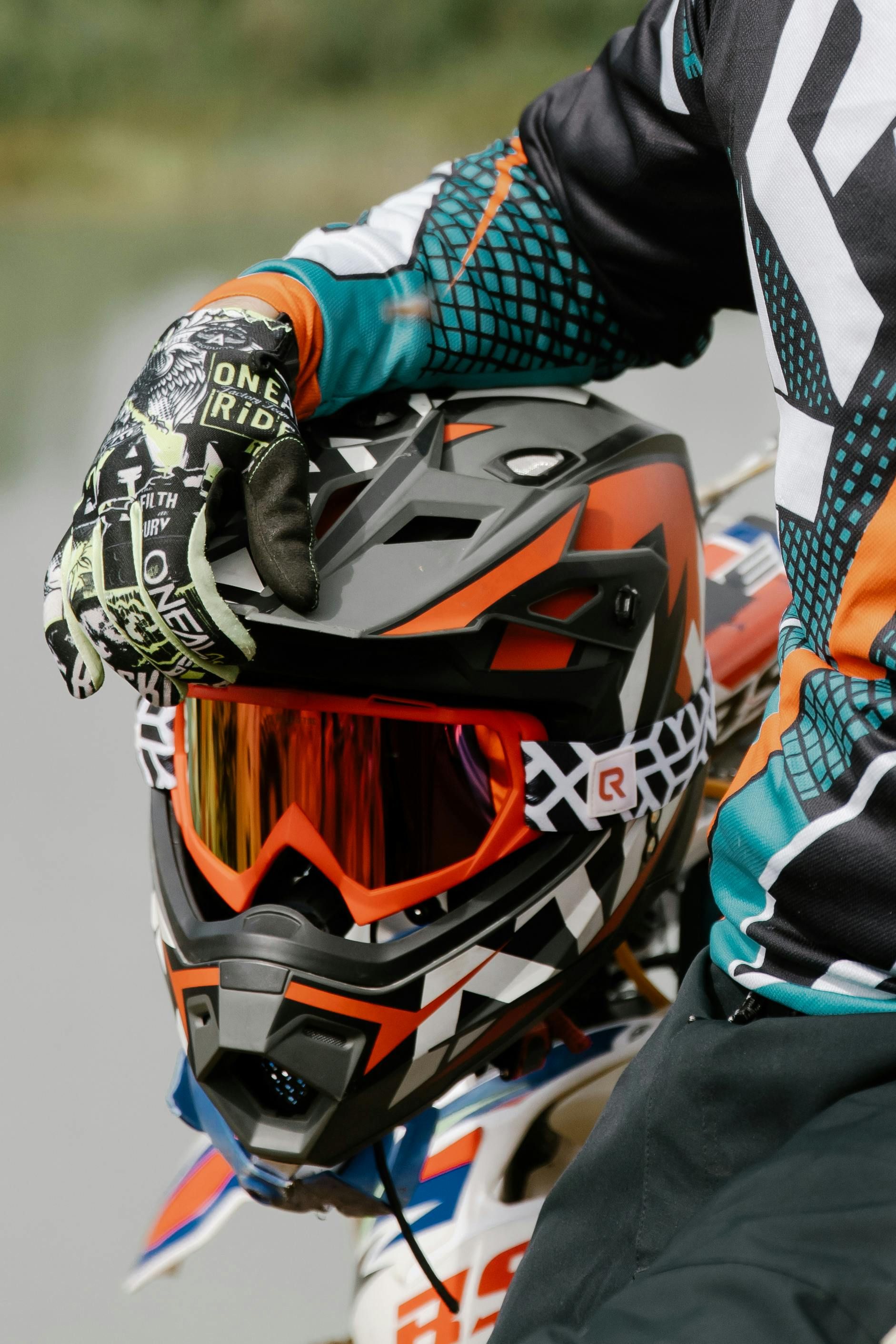 Motocross rider adjusting helmet with orange-tinted visor, wearing black, orange, and white gear.