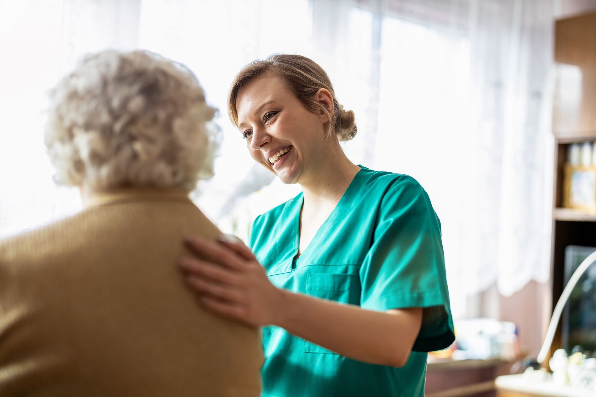 Smiling caregiver in green scrubs with hand on the shoulder of an elderly woman.