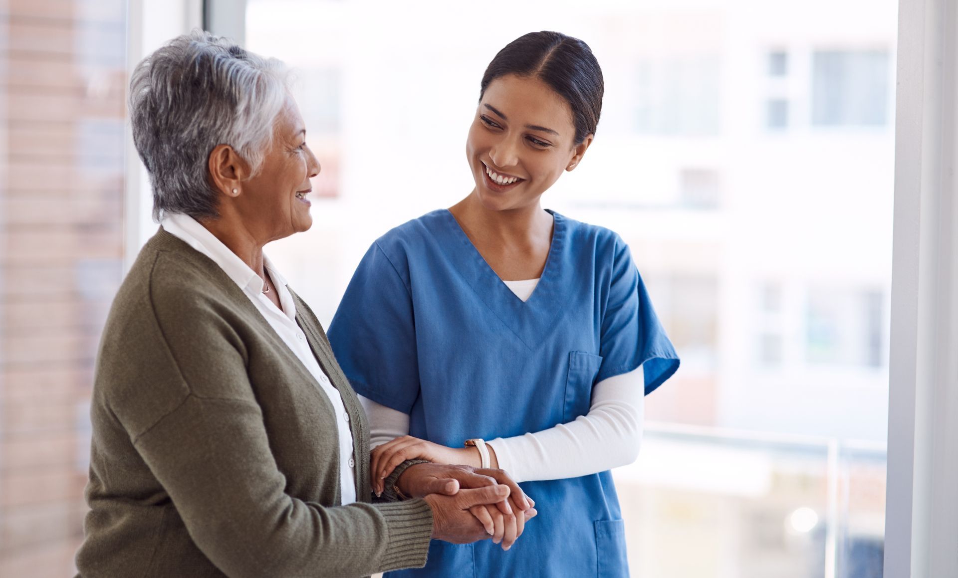 Nurse in blue scrubs assists an elderly woman. They smile, holding hands, near a window.