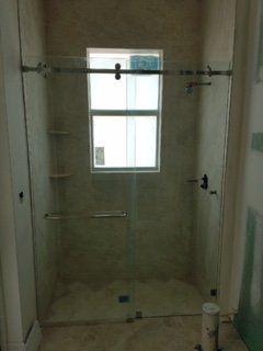 Empty tiled shower with glass door and a window, featuring a grab bar and wall-mounted shower fixtures.