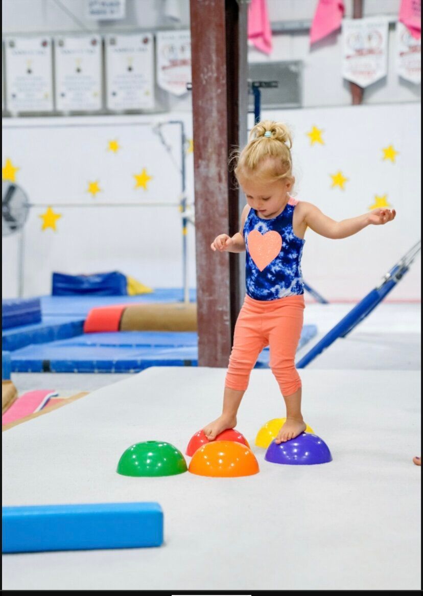 A little girl in a blue tank top is standing on a balance beam in a gym.