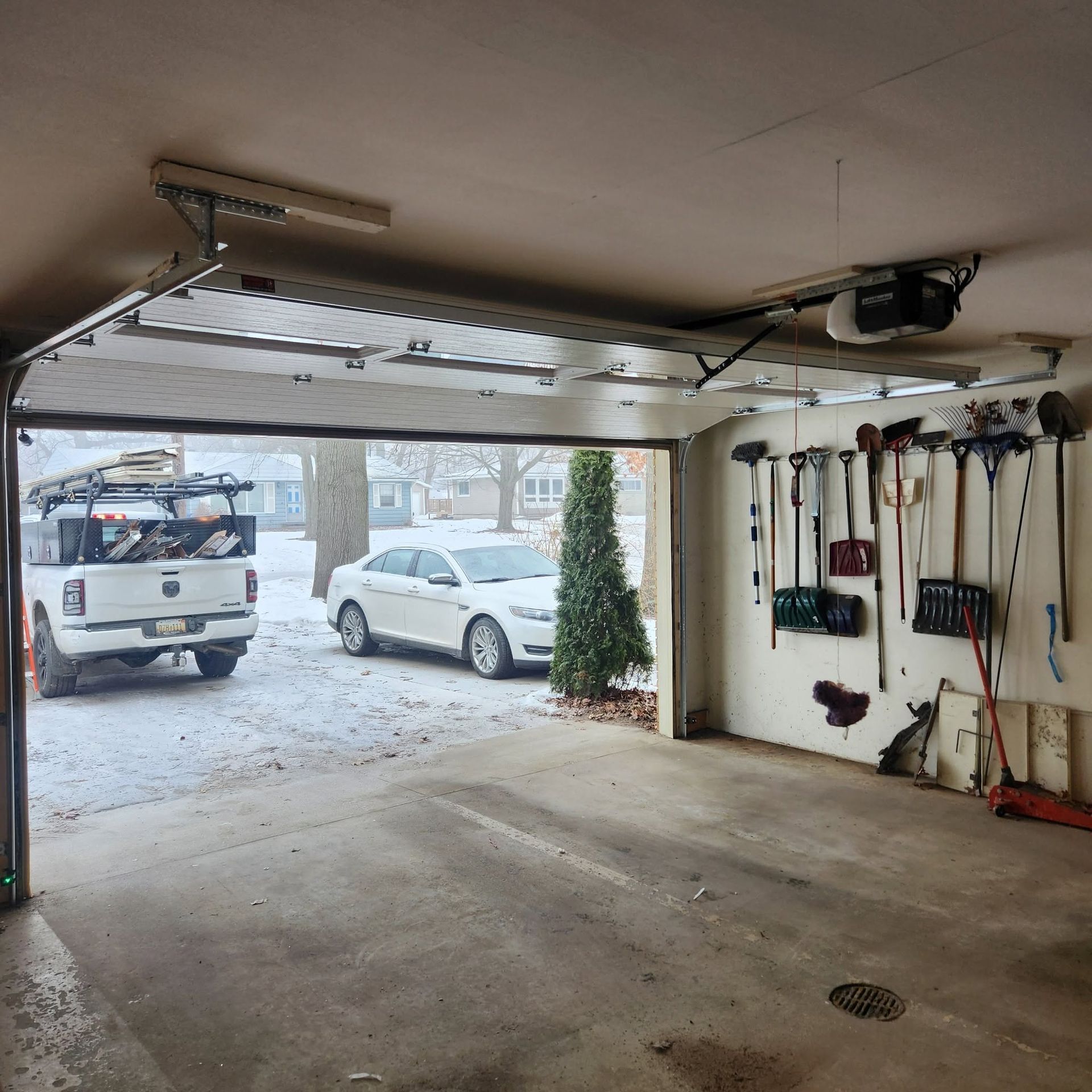 Snowy garage interior with an open door, tools on the wall, and two vehicles visible outside.