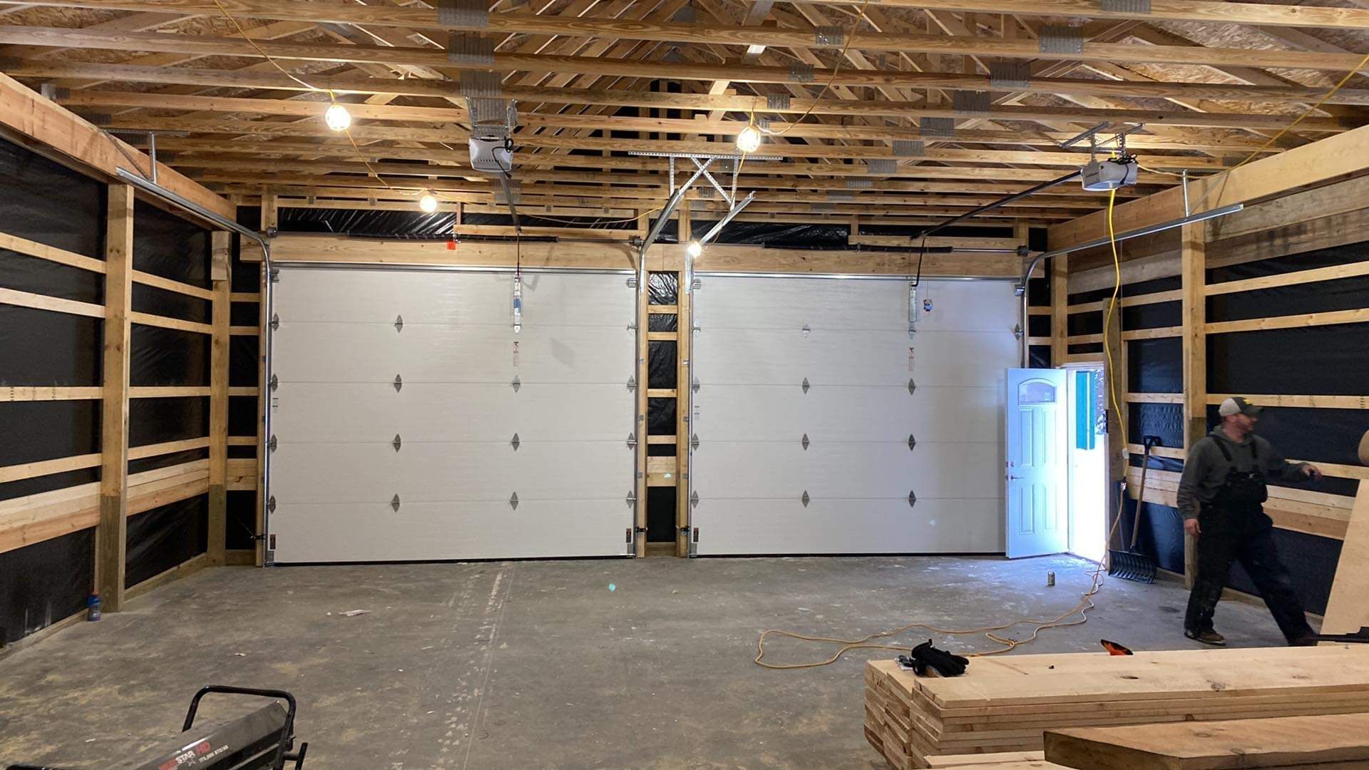 Empty warehouse interior with exposed wood framing, two large white garage doors, and a person working on the right