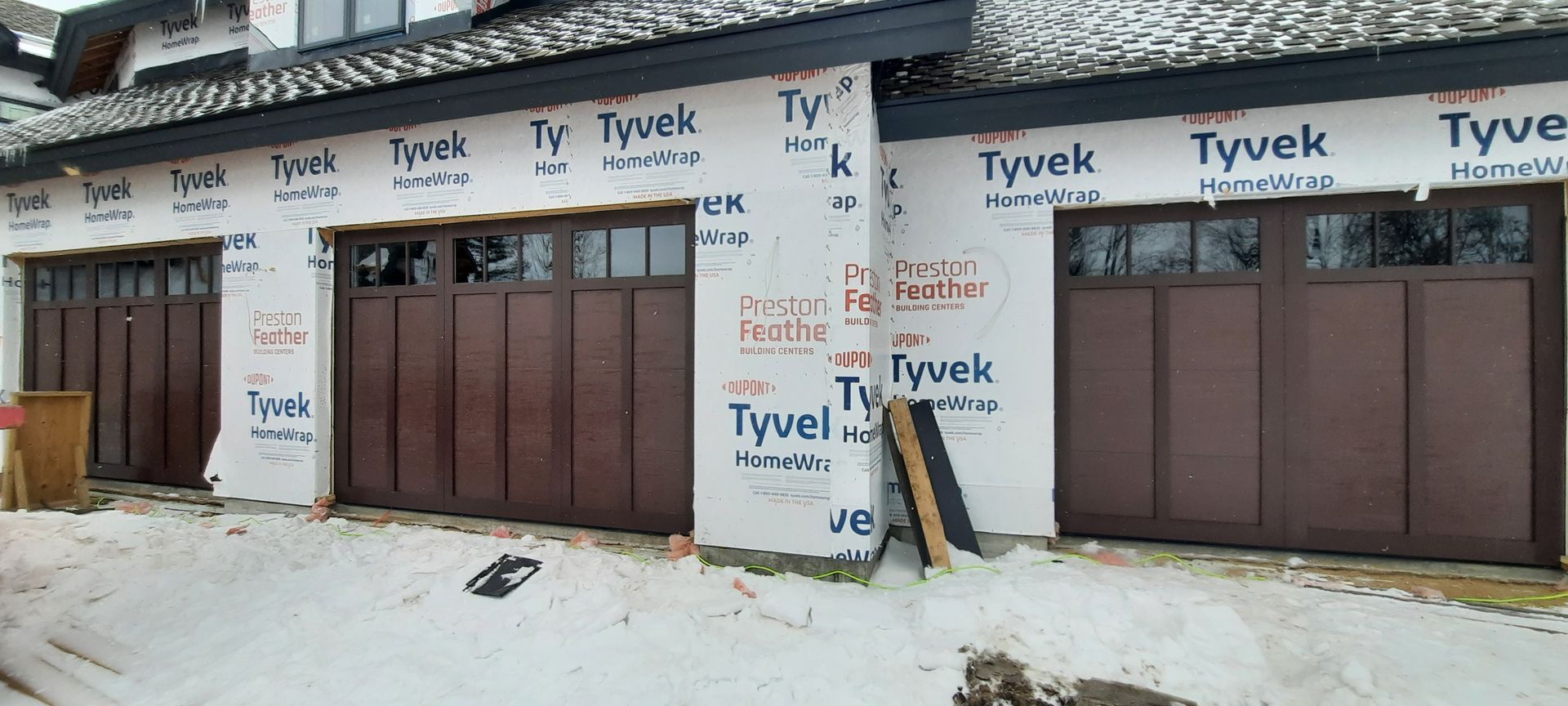Two unfinished townhouse garages wrapped in Tyvek, with brown doors and snow on the ground.
