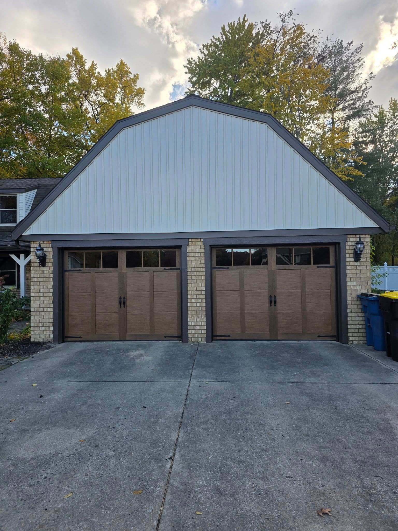 Two-car garage with white siding, brown doors, and a concrete driveway under trees.