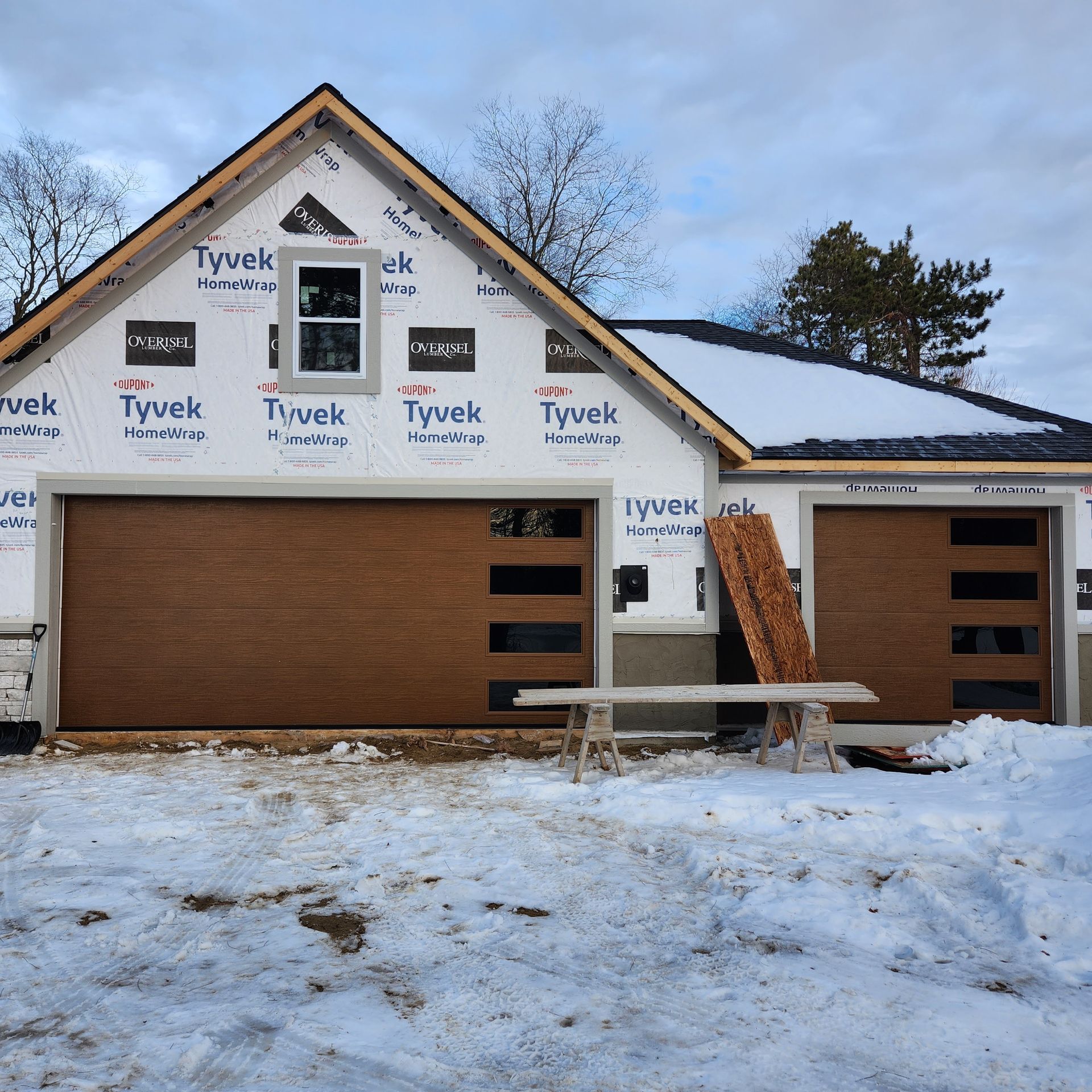 Snow-covered house under construction with brown garage doors and Tyvek-wrapped walls.