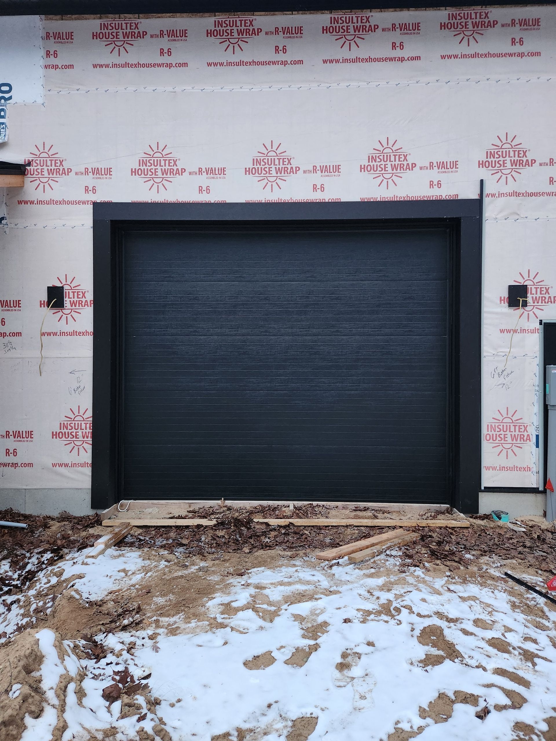 Closed black garage door in an unfinished building with snow and debris in front.
