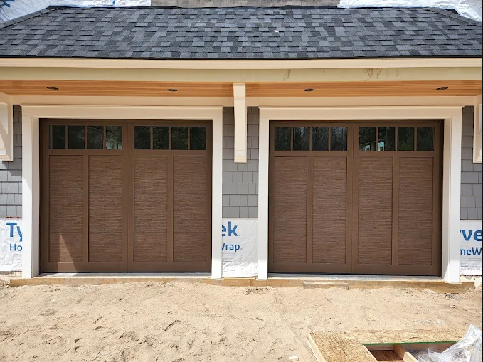 Two brown garage doors on a house under construction, with white trim and unfinished ground.