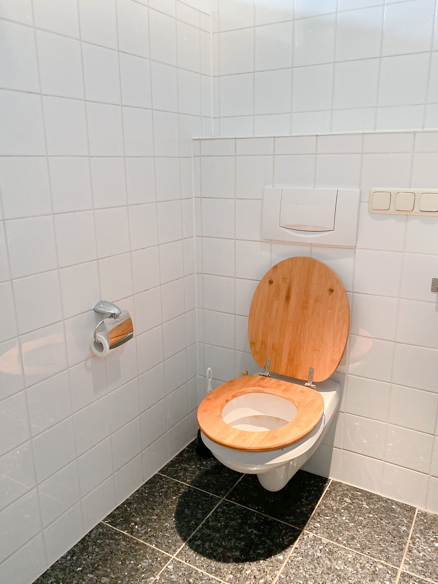 A toilet with a wooden seat is in a bathroom with white tiles.