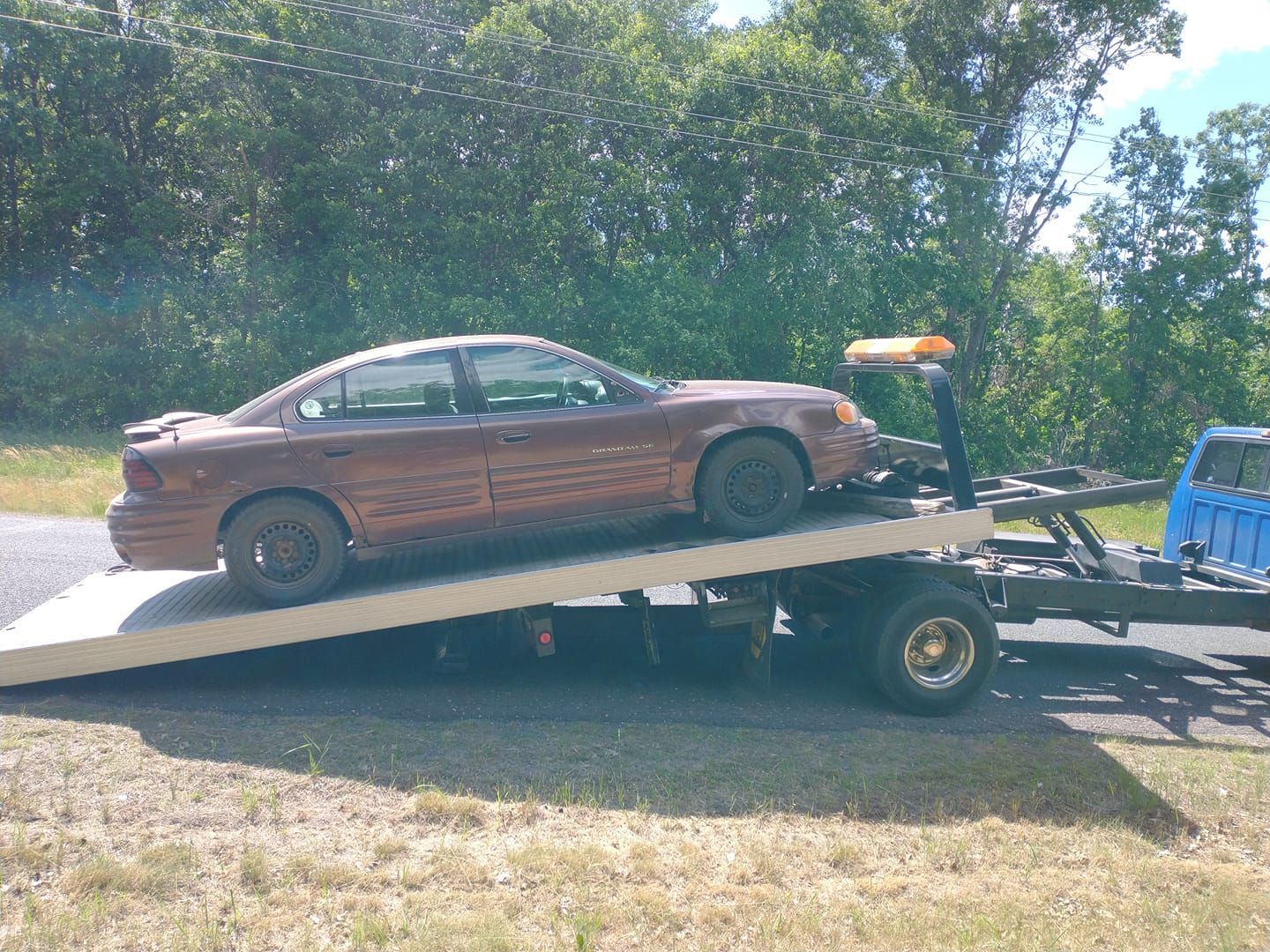 Brown sedan on a flatbed tow truck. Sunny outdoor setting with trees in the background.