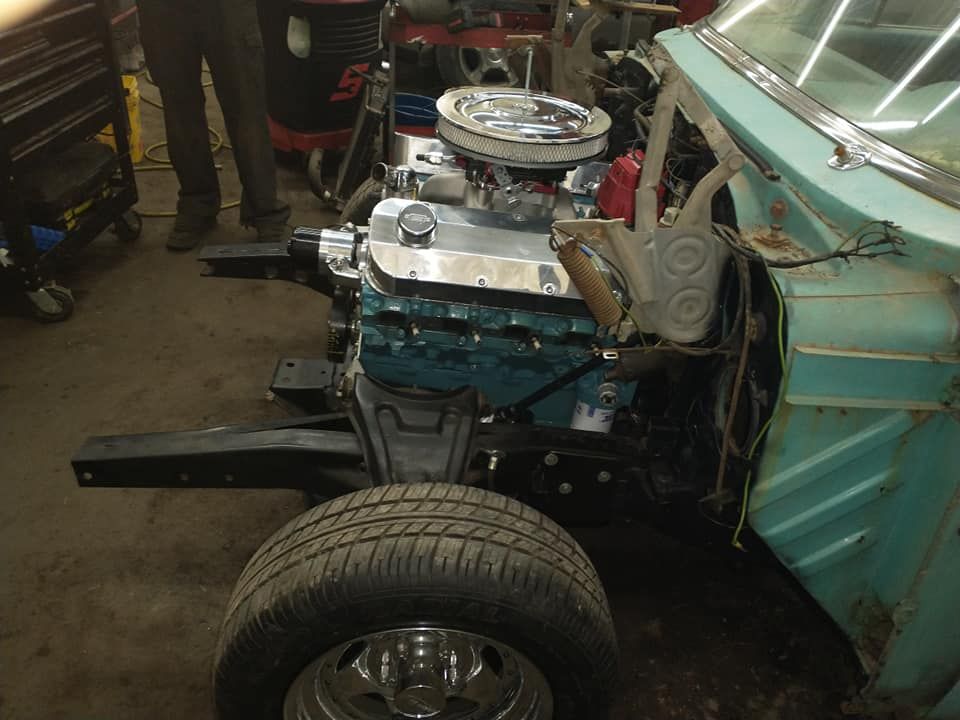 Classic car chassis with engine installed, front tire. Teal body panels, black frame. Garage setting.