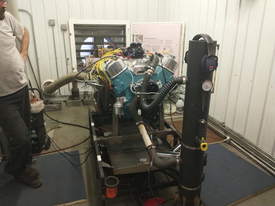 Engine dynamometer test cell; engine is teal, on a metal frame. Person observing.