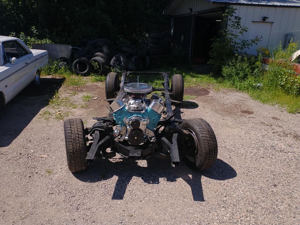 Car chassis with blue engine, tires, and frame on gravel, near a building and car.
